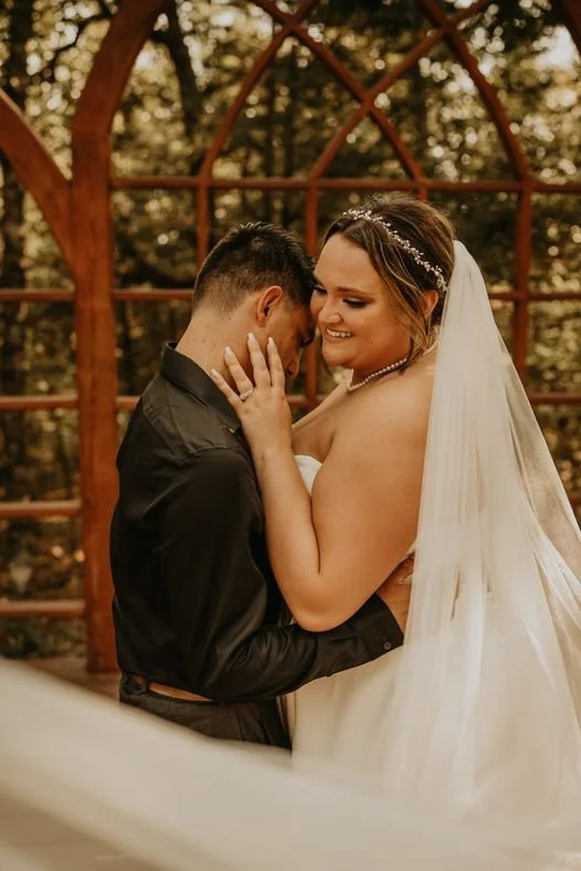 A bride and groom embrace during their wedding ceremony outdoors, with a wooden arbor and trees in the background.