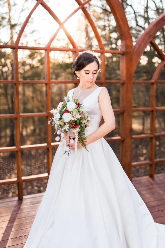 A bride in a white wedding dress holding a bouquet of flowers, standing outdoors on a wooden platform with a wooden arch and trees in the background during sunset.