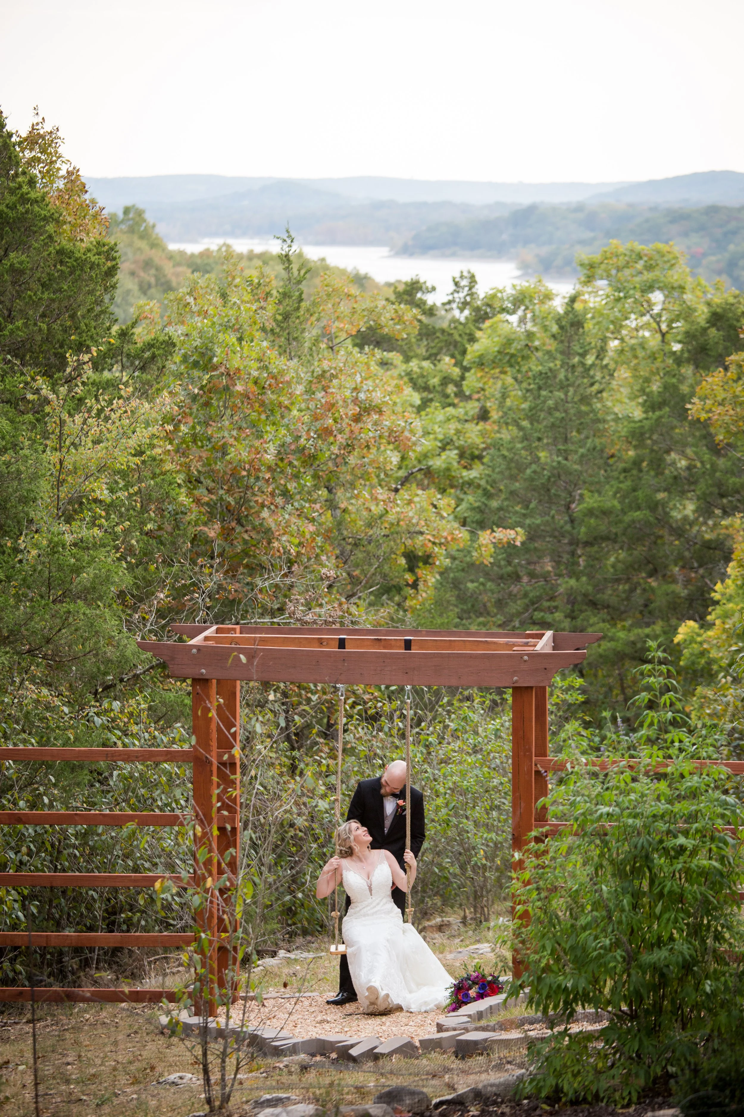 A bride in a white wedding dress sitting on a swing with a groom standing behind her, holding the swing chain, in a lush outdoor setting with trees and a river lake in the background.
