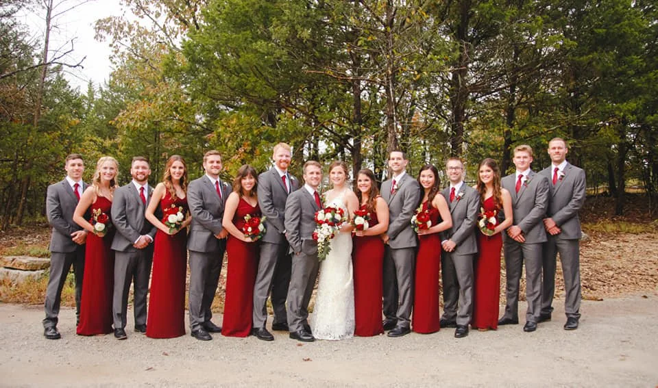 A wedding party of 14 people, including the bride and groom, standing outdoors in front of trees. The women are in red dresses, holding bouquets, and the men are in gray suits with red ties.
