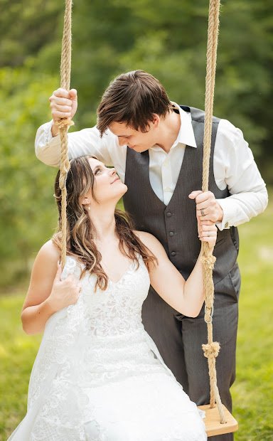 A woman in a white wedding dress and a man in a vest and white shirt standing on a swing, looking at each other affectionately outdoors in a park or garden.