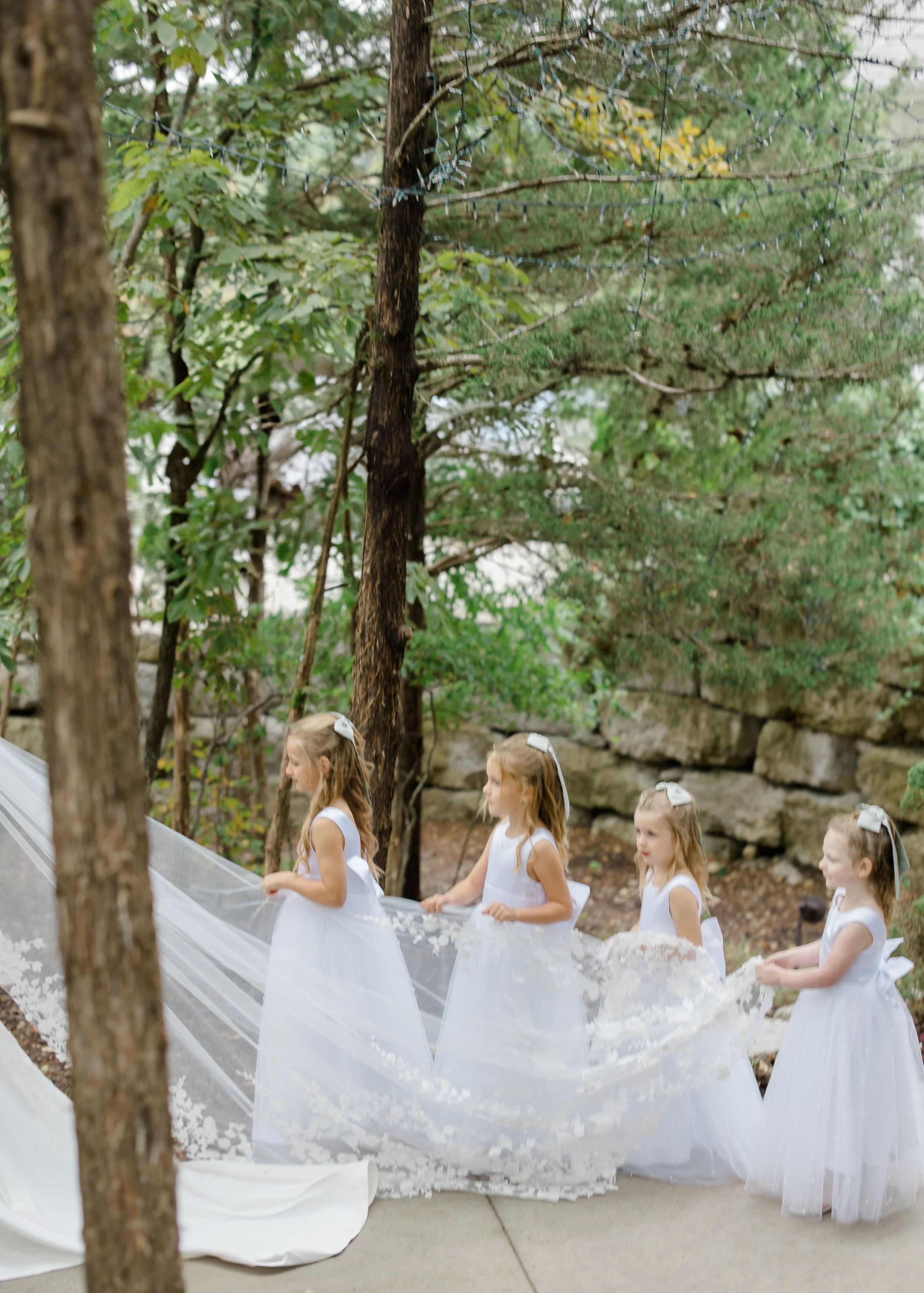 Four young girls in white dresses participating in a procession outdoors, holding a long white fabric ribbon with floral accents, with trees and a stone wall in the background.