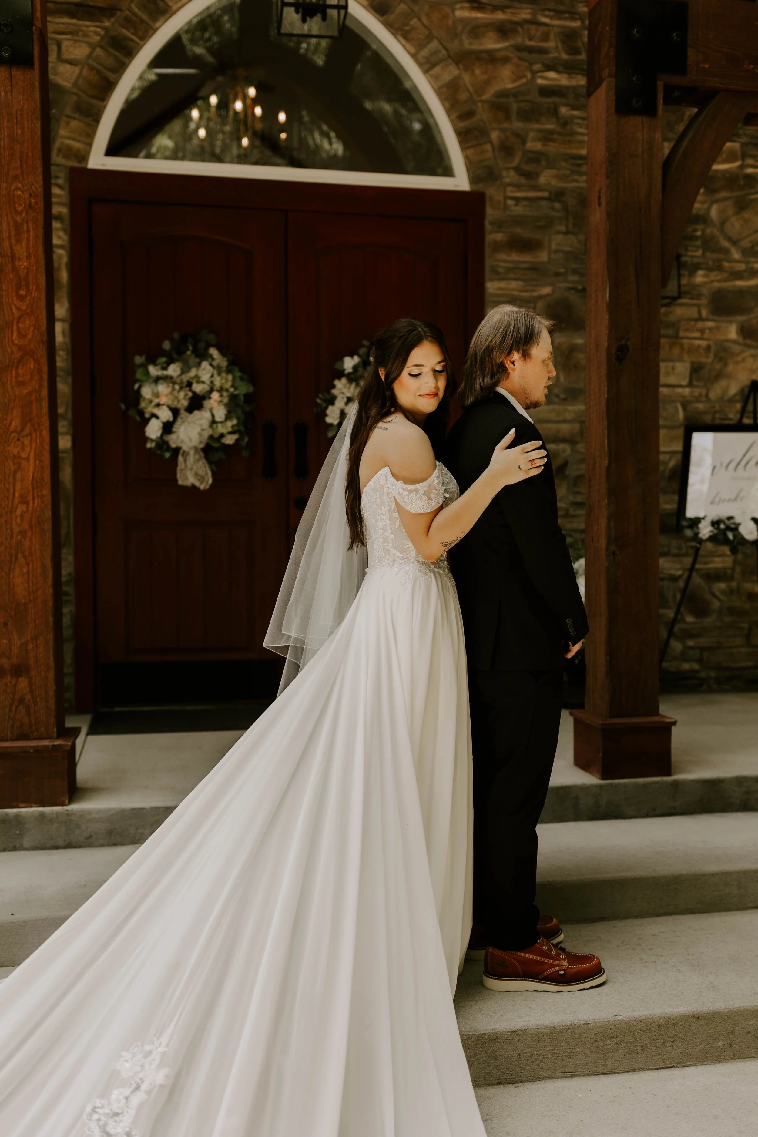 A bride in a white wedding gown with lace details on the shoulders, holding onto a groom dressed in a black suit. They are standing inside a rustic venue with a stone wall, wooden beams, and floral decorations, during a wedding ceremony.