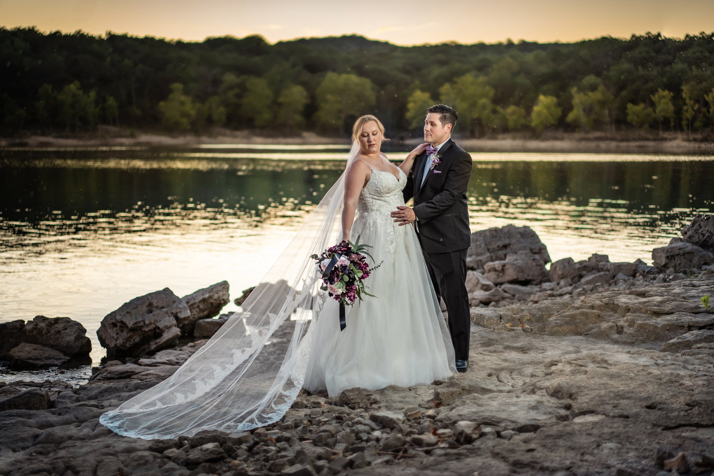 A bride and groom standing on rocks by a body of water during sunset, with the bride holding a bouquet and wearing a wedding gown, and the groom dressed in a suit with a bow tie.