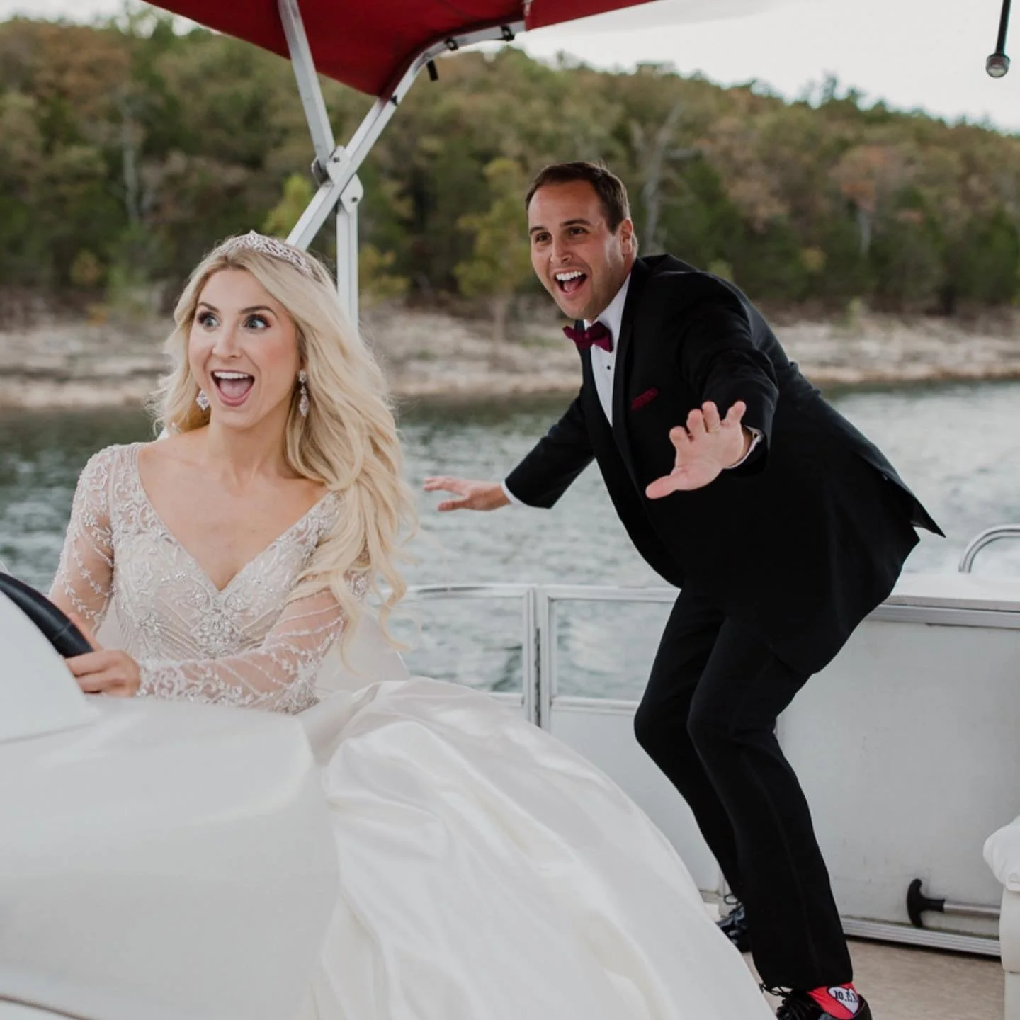 A bride and groom on a boat, with the bride driving and the groom jokingly acting as if he is falling or losing balance, both smiling and having fun.
