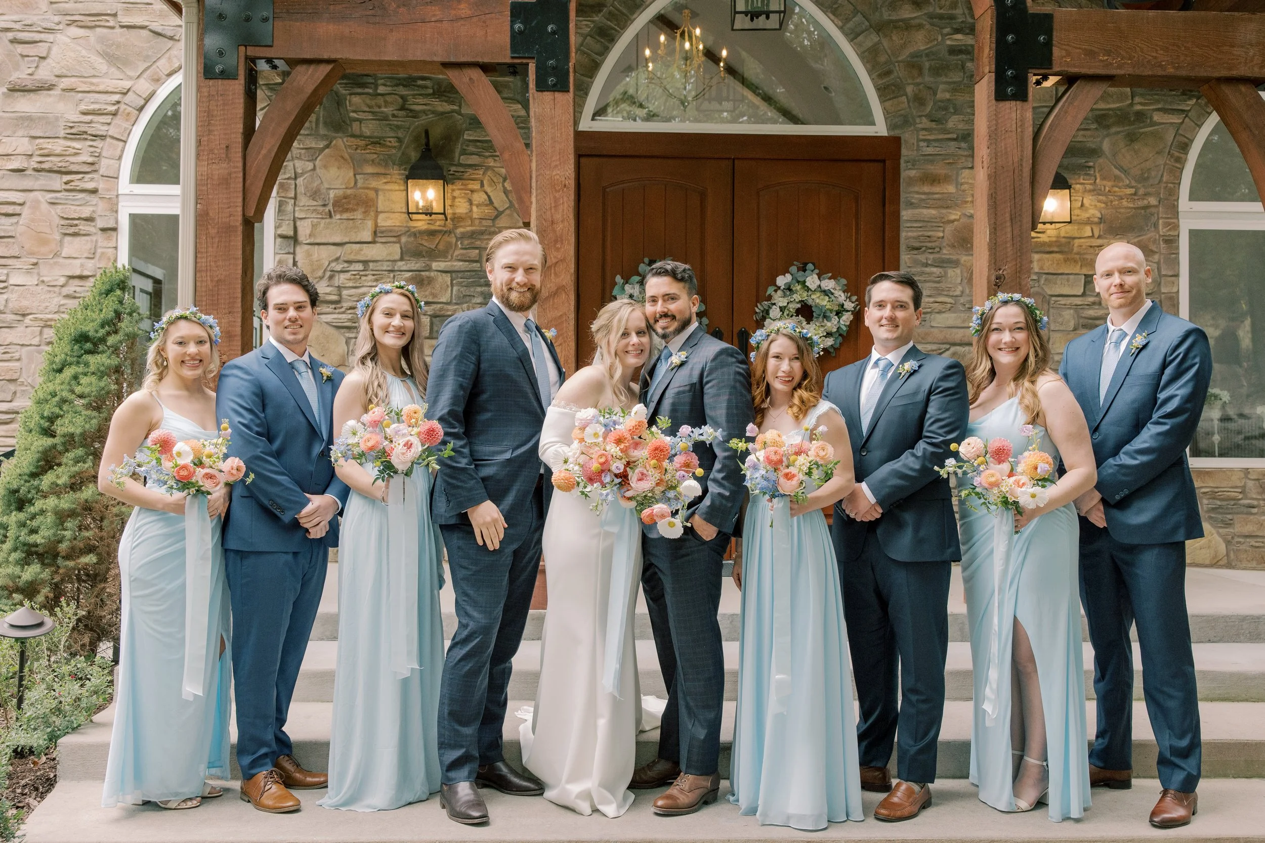 A group of nine people, including a bride and groom, standing outside a stone building with wooden beams and arched windows, dressed in wedding attire, holding bouquets of flowers, smiling for a wedding photo.