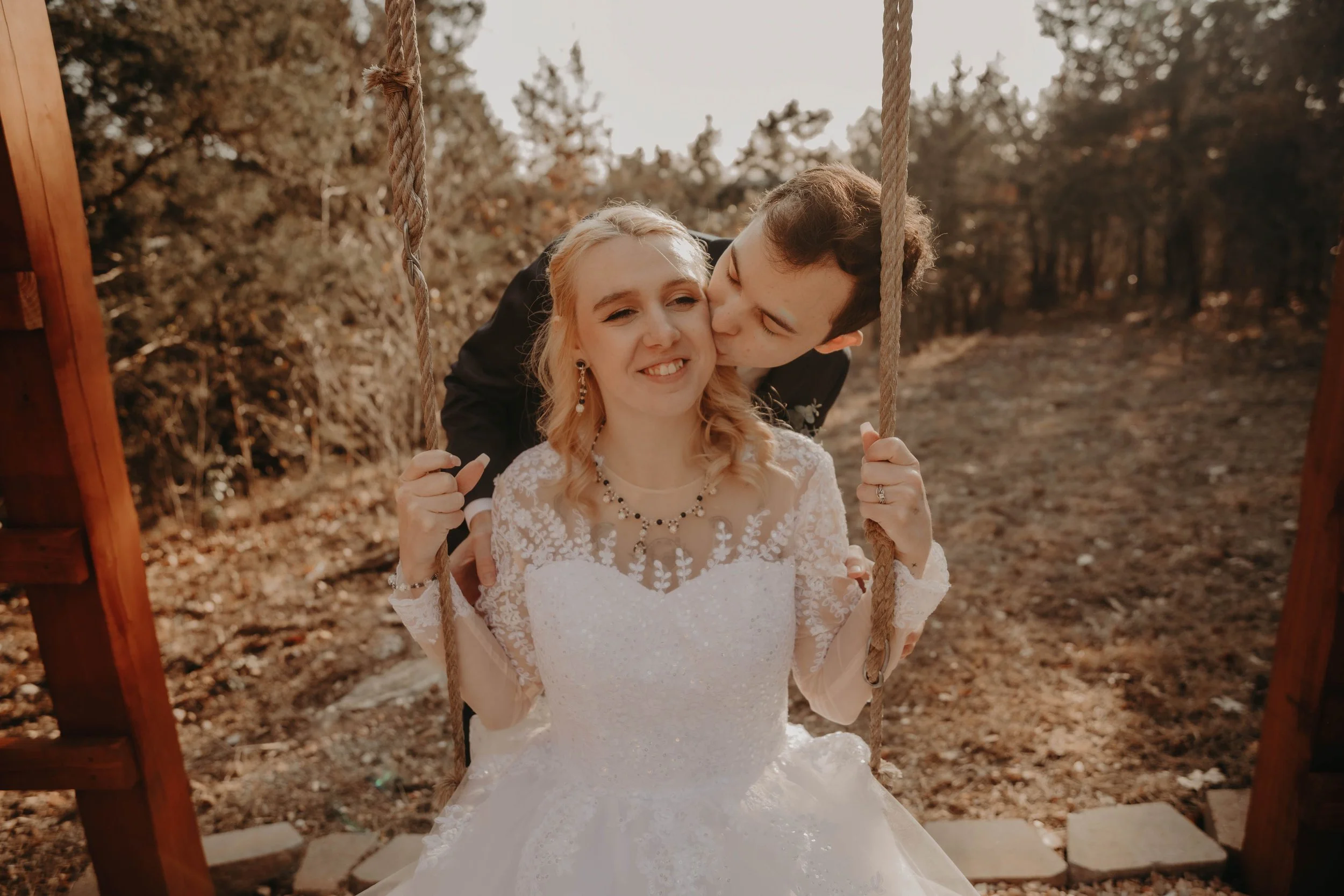 A bride with blonde hair seated on a swing, being kissed on her cheek by a groom with dark hair, outdoors with trees and fallen leaves in the background.