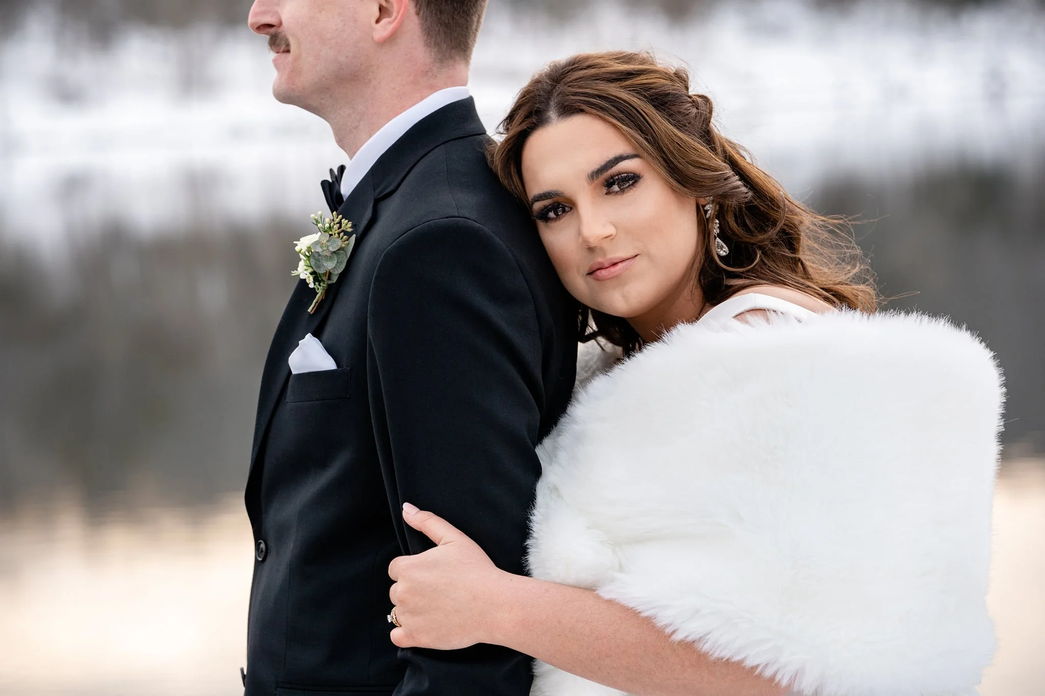 A bride resting her head on a groom's shoulder outdoors in winter, both dressed formally. The bride has dark hair and wears earrings and a white fur wrap, while the groom is in a black tuxedo with a boutonnière.
