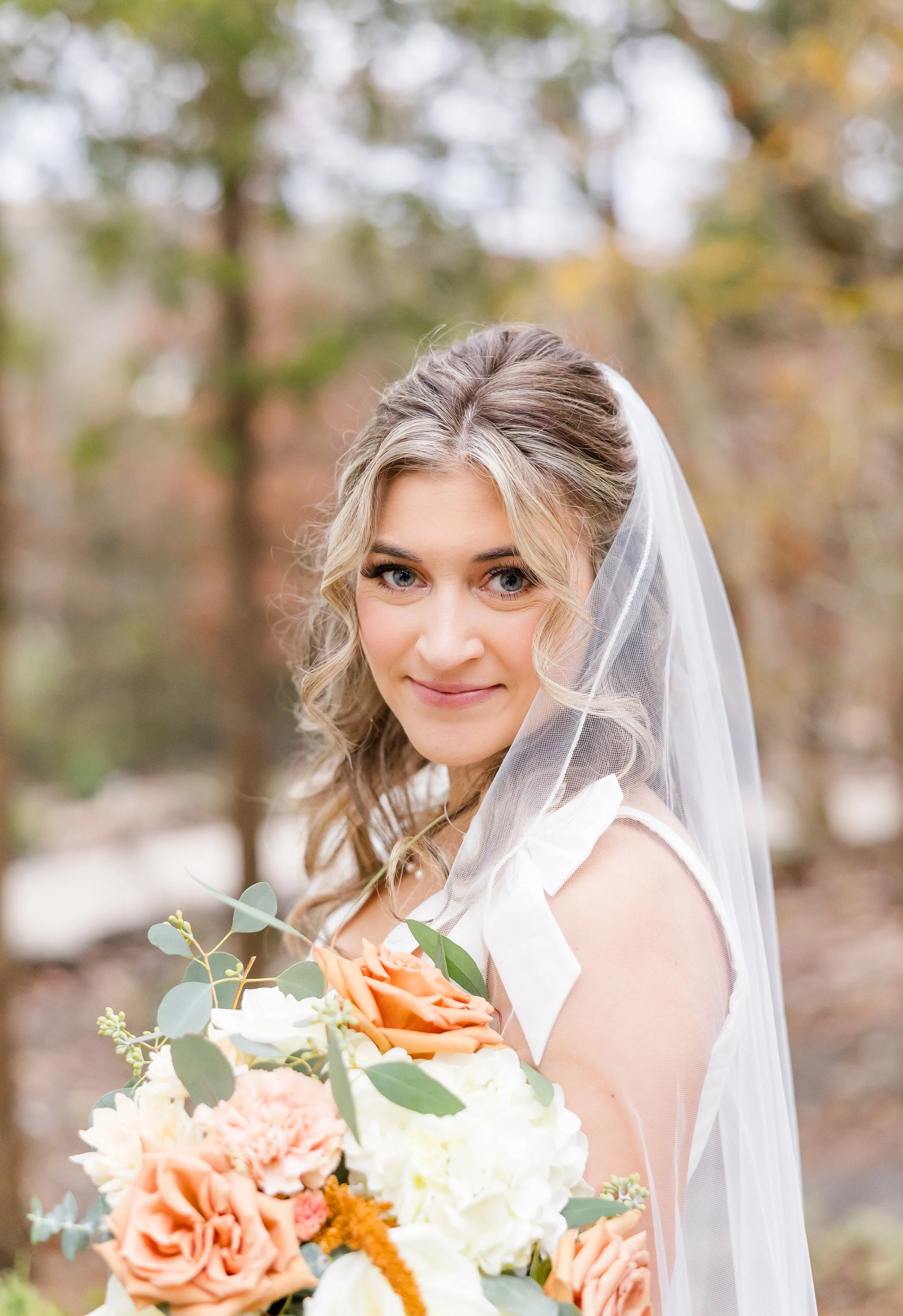 A bride with blonde hair styled in loose waves, wearing a white wedding dress and veil, holding a bouquet of orange, white, and green flowers outdoors with autumn trees in the background.