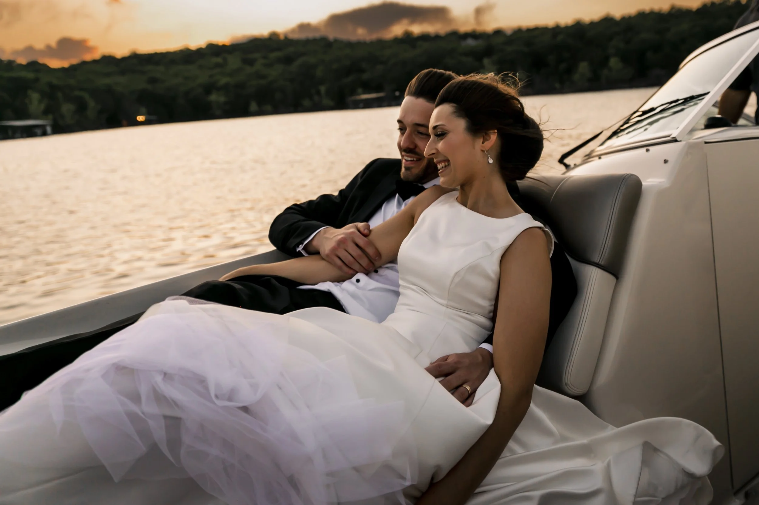 A couple in formal attire is sitting on a boat enjoying a sunset on the water. The woman is wearing a white dress and earrings, and the man is wearing a tuxedo. They are smiling and looking at each other, with the woman's head resting on the man's sh