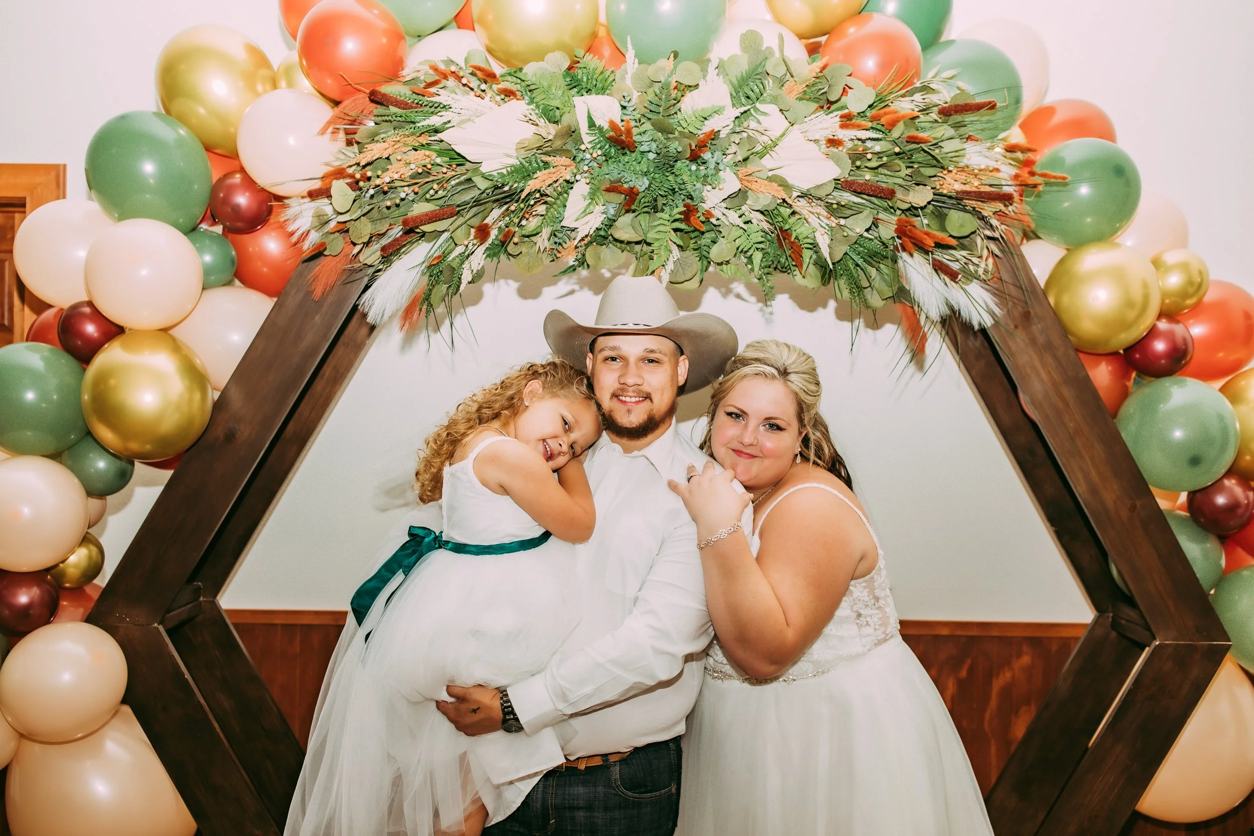A family of three, including a man, woman, and young girl, posing under a decorative arch of balloons and floral arrangements at a celebration or wedding event.