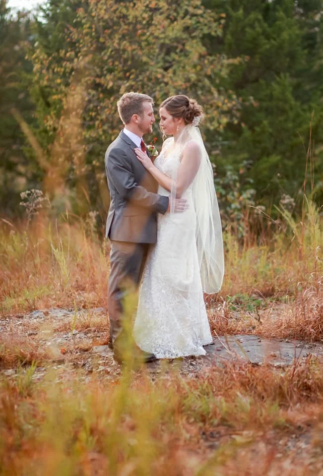 A bride and groom standing close together outdoors, facing each other with smiles, in a natural setting with trees and grass.
