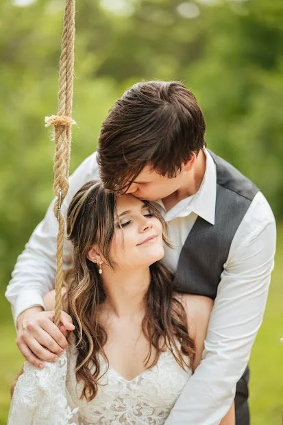 A young couple sharing a romantic moment on a swing outdoors, with the man kissing the woman's forehead. Green trees in the background.