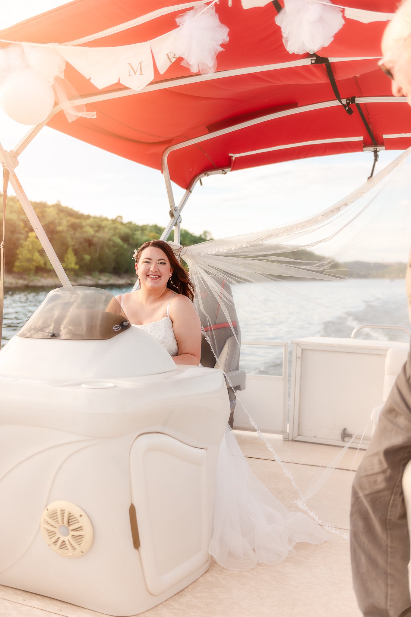 A smiling bride sitting on a boat with white angel wings wearing a wedding dress. The boat has a red canopy decorated with white tulle and balloons, and the background features a river and trees.