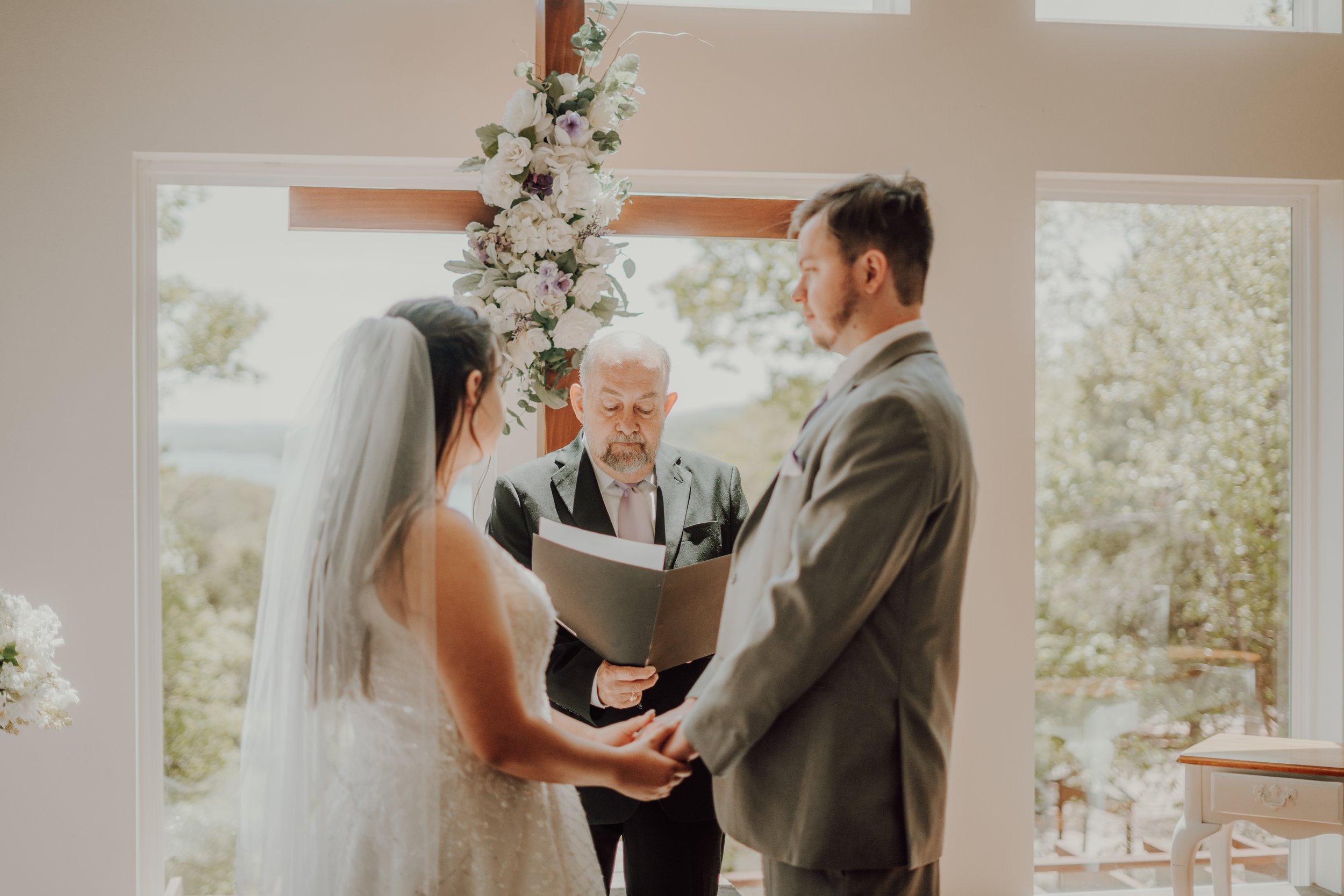 A couple getting married exchanging vows in front of a cross decorated with white and purple flowers during their wedding ceremony.