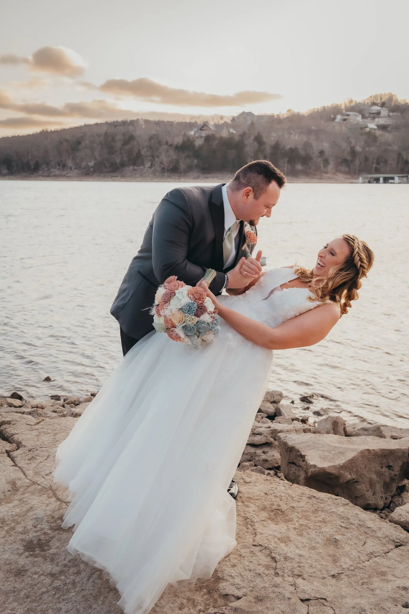 A newlywed couple is by a lake on a rocky shore, with the groom dipping the bride in a dance pose, both smiling joyfully. The bride is holding a bouquet of pastel roses, and the background features a hillside with houses and a partly cloudy sky.
