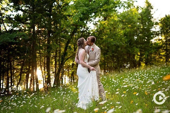A bride and groom kissing in a grassy field with wildflowers, trees in the background, during sunset.