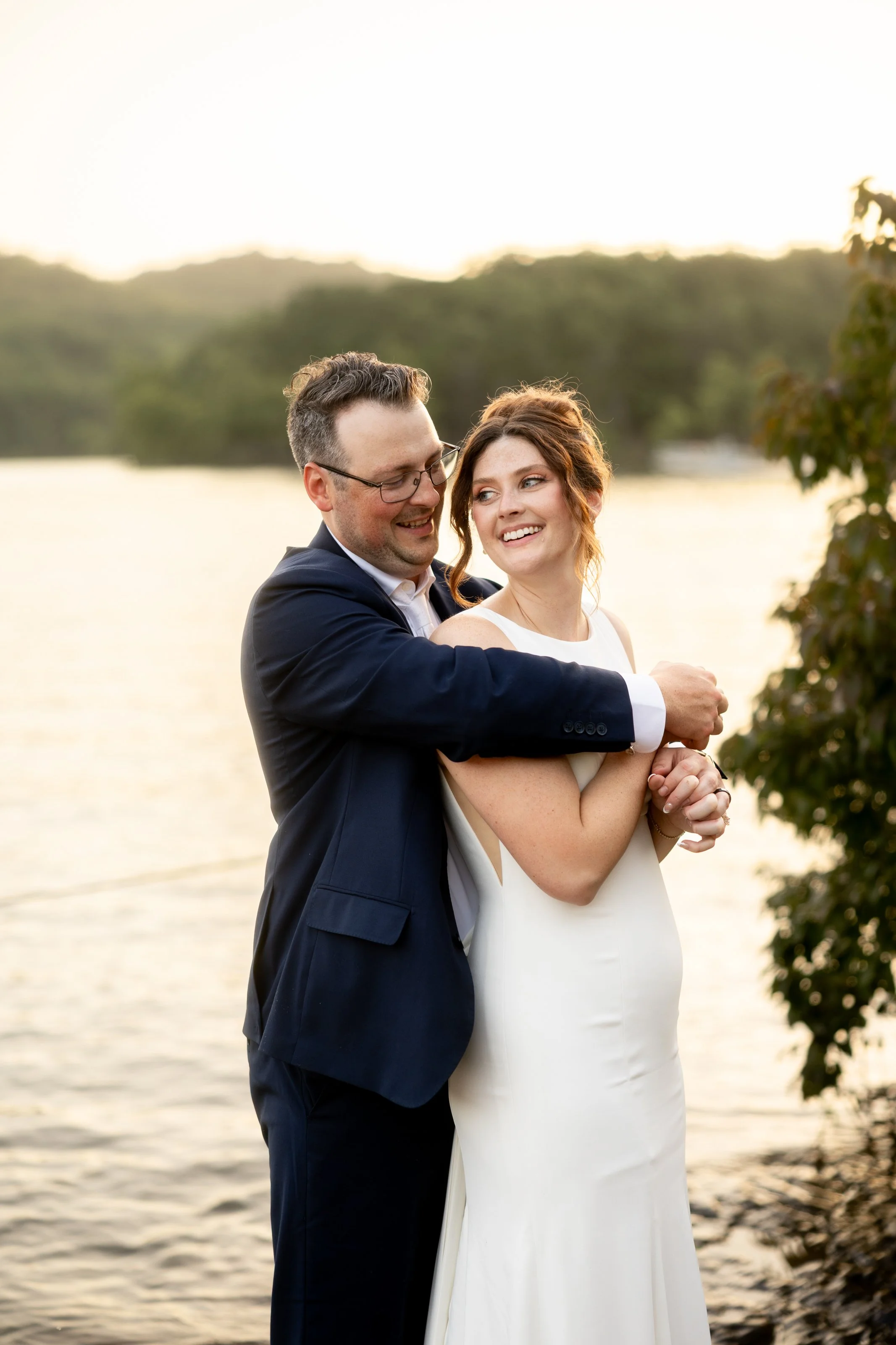A happy couple in wedding attire standing near a lake during sunset, with the man embracing the woman from behind.