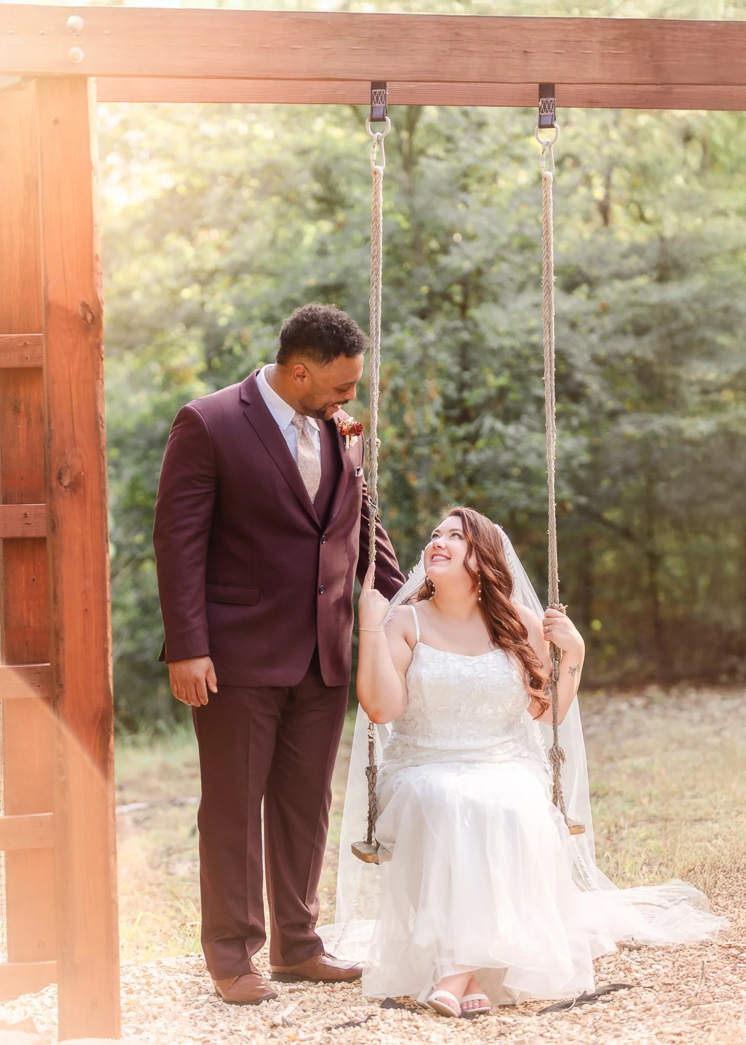 A newlywed couple, a man in a dark suit and a woman in a white wedding dress sitting on a swing outdoors, with the man looking fondly at the woman who is smiling at him. The background features green trees and soft sunlight.