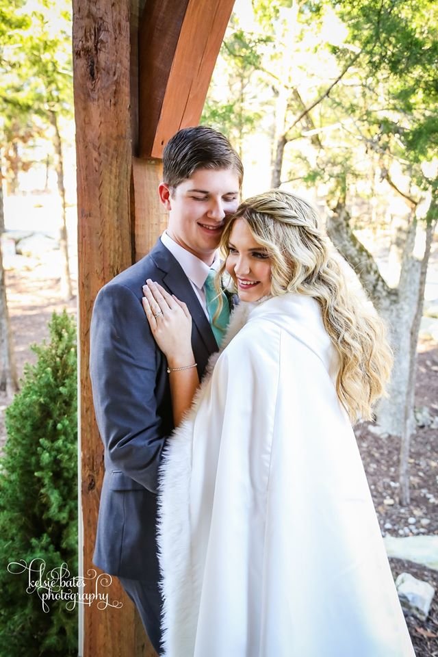 A couple dressed in formal attire, standing closely together outdoors during daytime, with trees and sunlight in the background.