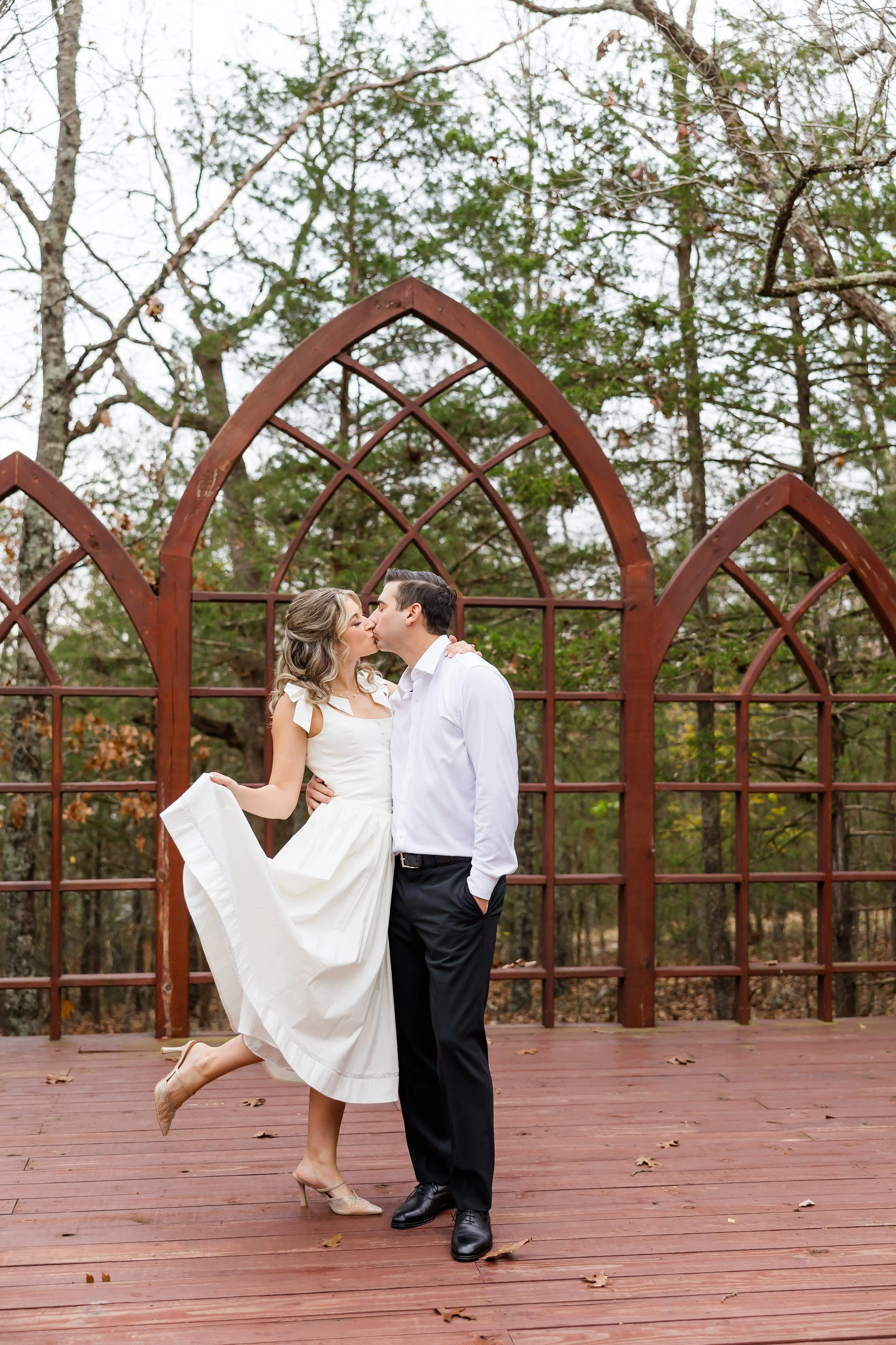A couple kissing outdoors on a wooden deck surrounded by trees, with a rusty metal arch structure in the background. The woman is wearing a white dress and heels, and the man is dressed in a white shirt and black pants.