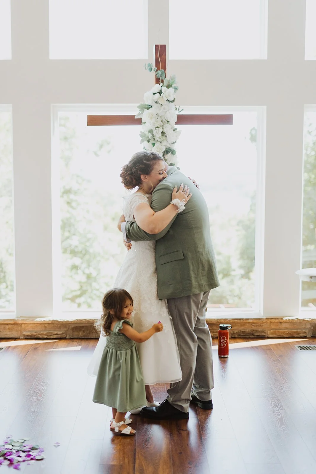 A bride and groom hugging in front of a large cross with white flowers at a wedding ceremony, with a little girl smiling nearby.