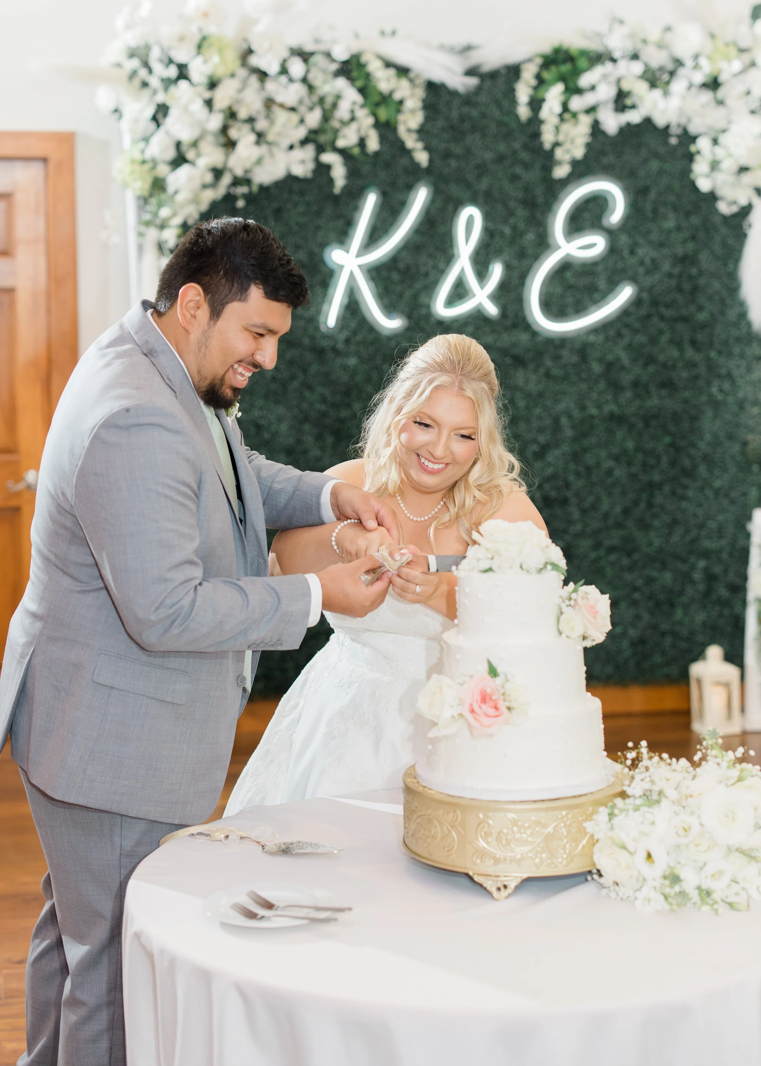 A bride and groom at a wedding cake-cutting ceremony, cutting into a three-tiered white wedding cake decorated with pink and white roses, with a green leafy backdrop and a neon sign displaying their initials 'K & E' behind them.