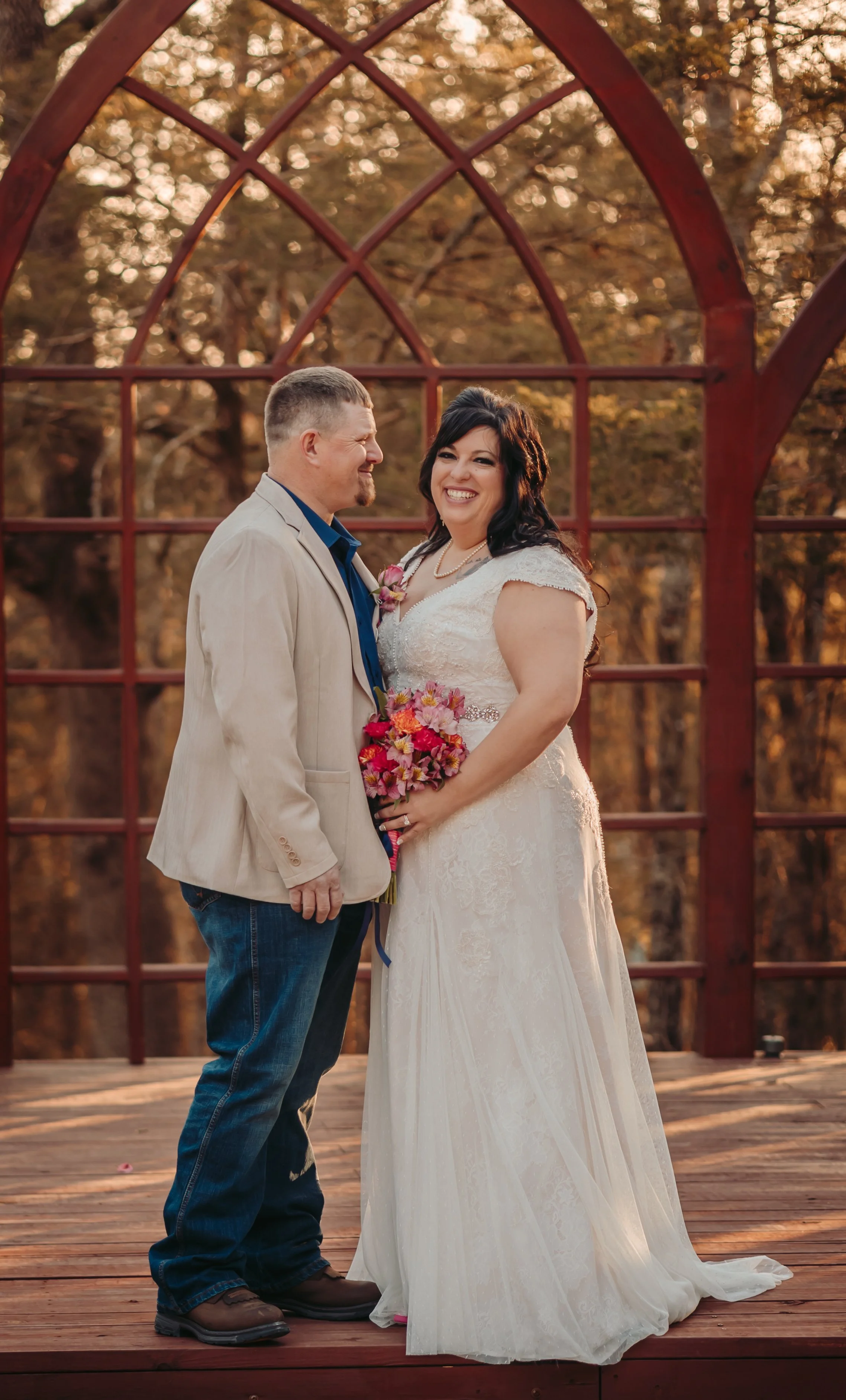 A bride and groom stand on a wooden deck with a red metal arch behind them, surrounded by autumn-colored trees, during sunset. The groom, with short hair and a light-colored suit jacket, is smiling as he looks at the bride. The bride, with long dark hair and a white lace wedding gown, is holding a bouquet of pink, purple, and orange flowers, smiling broadly at the camera.