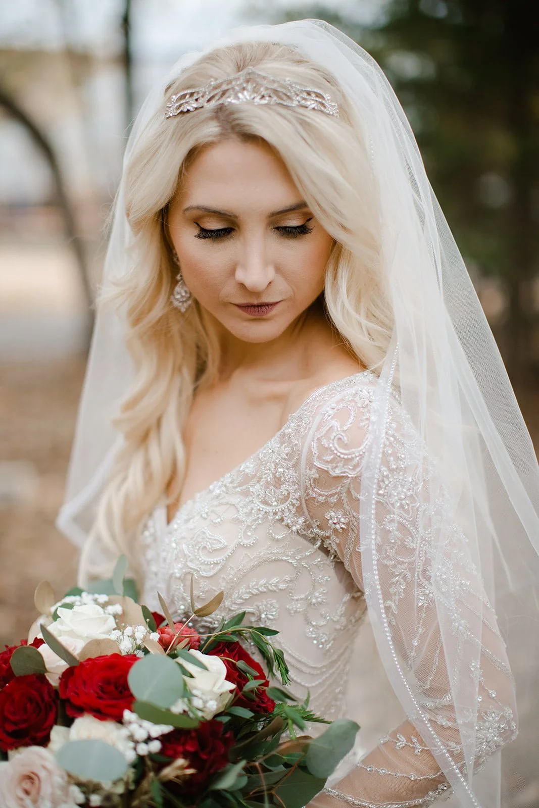 A bride in a wedding dress with lace and bead details, holding a bouquet of red, white, and pink flowers with green foliage, outdoors with blurred trees in the background.