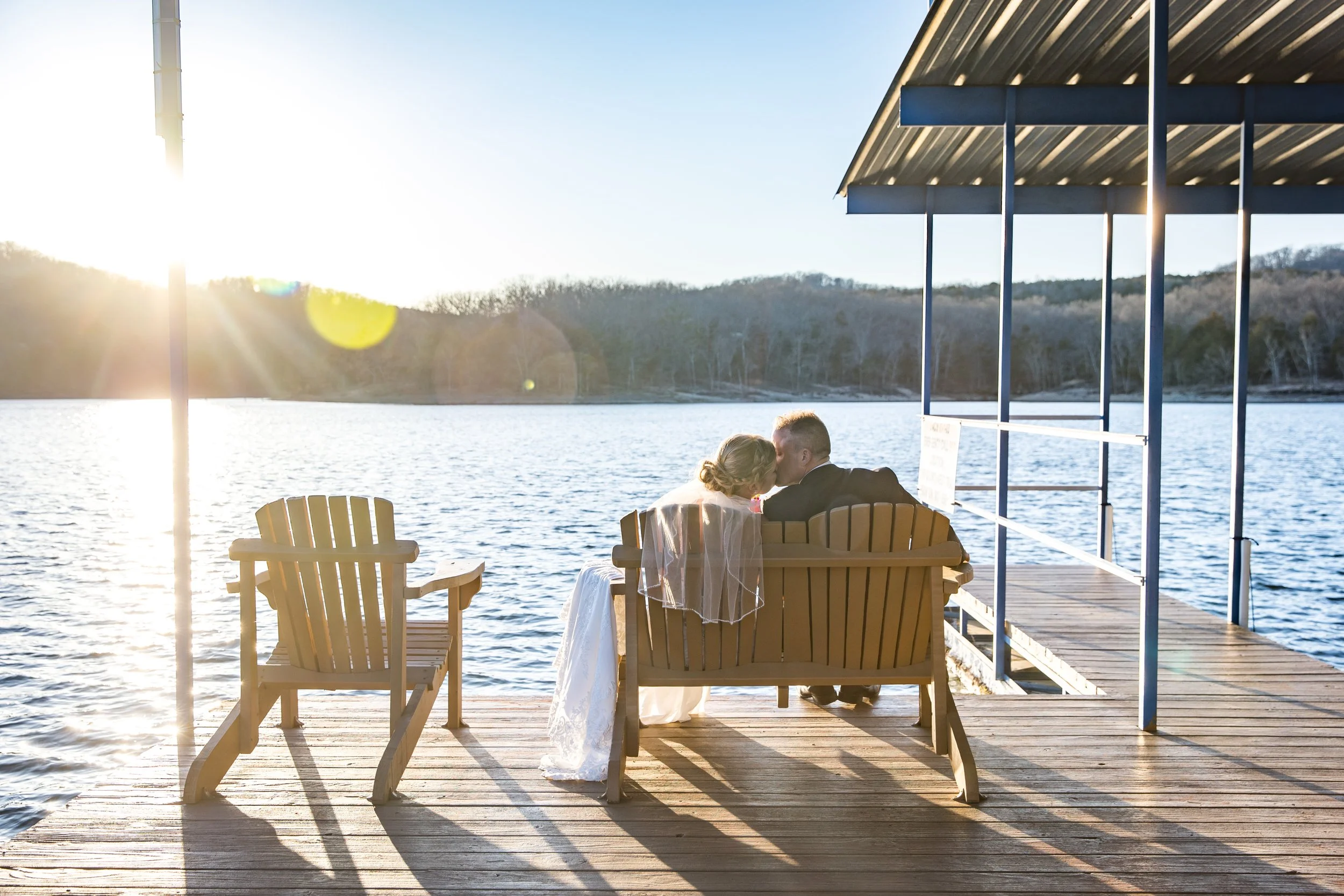 A couple kissing on a dock by a lake during sunset, with empty wooden chairs and a boat dock in the background.
