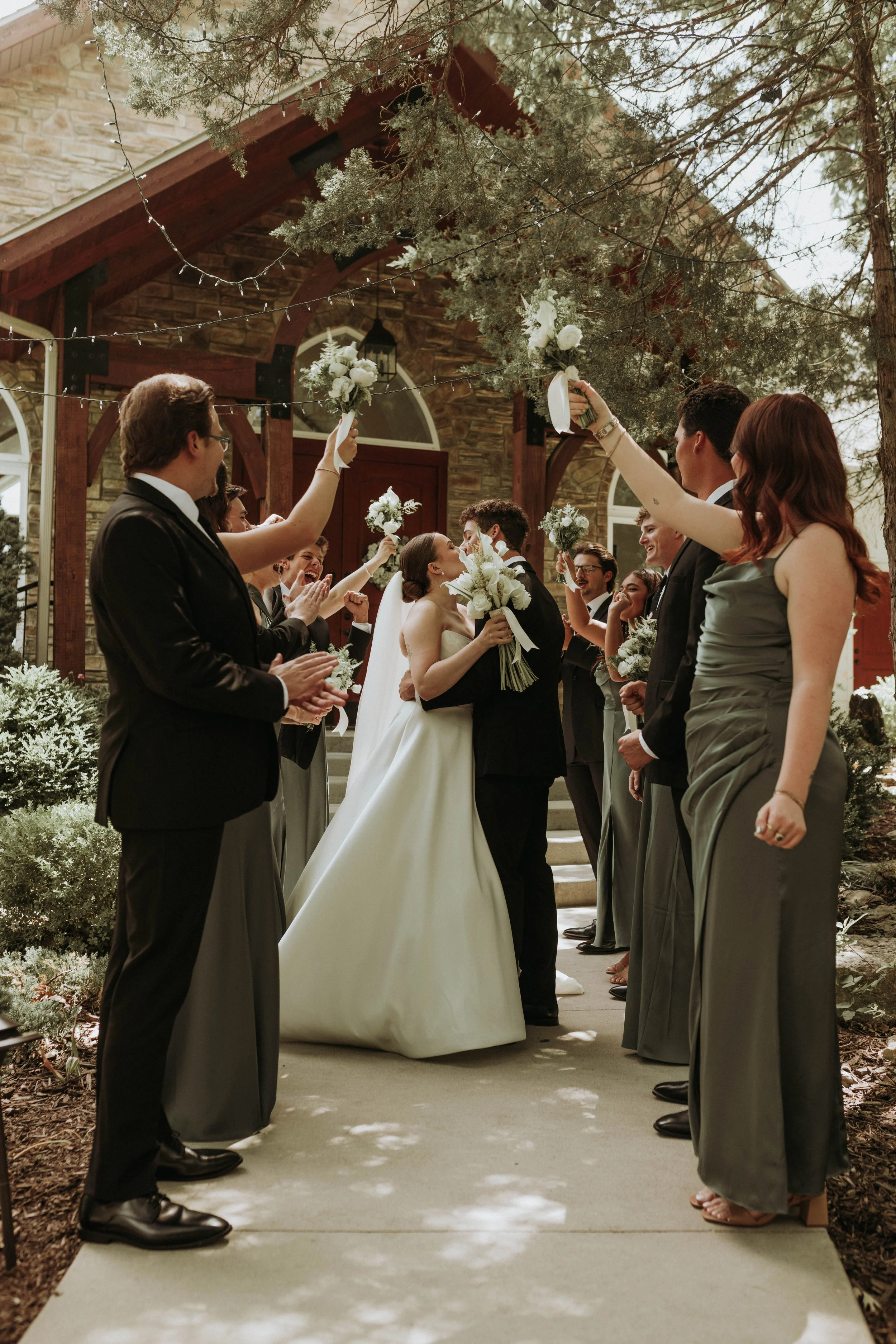 Bride and groom kissing under hanging floral arrangements and surrounded by friends during a wedding ceremony outside of a house, with guests holding bouquets and clapping.