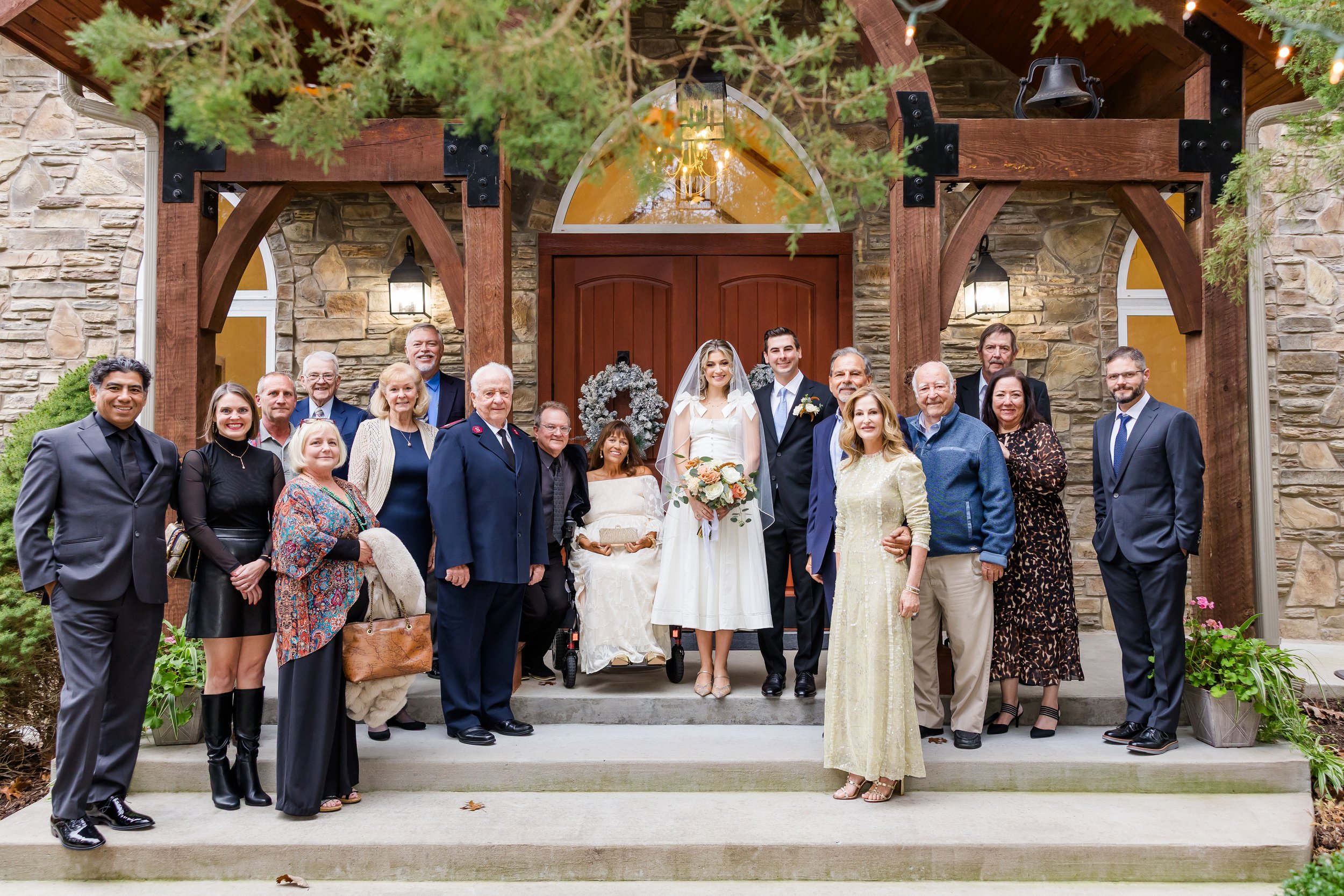 Group of people, including a bride in white dress and veil, and groom in suit, standing with family and friends on steps outside of a stone house, celebrating a wedding.