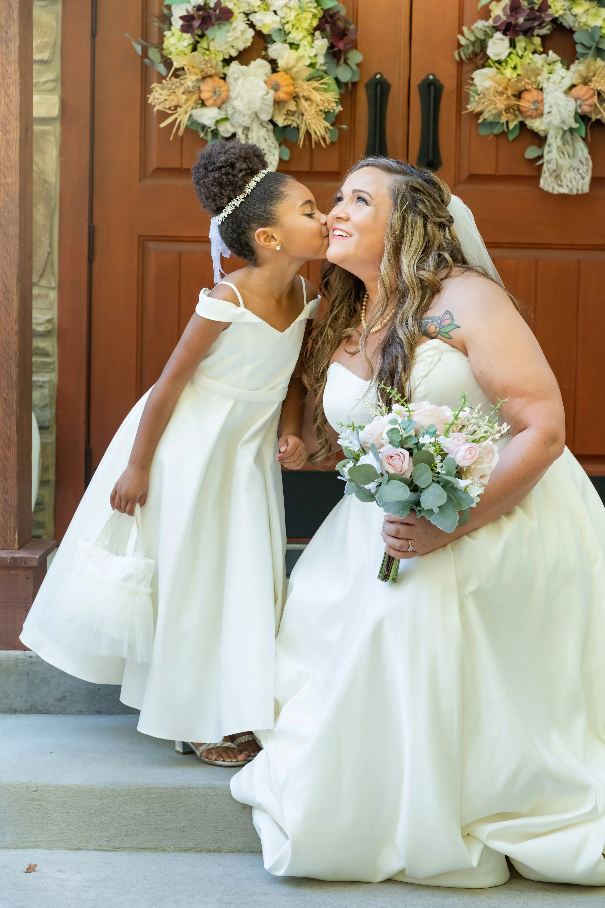 A bride in a wedding gown holding a bouquet of pink roses and greenery, kneeling on a concrete step, as a young girl kisses her on the cheek in front of a wooden door decorated with fall-themed wreaths.
