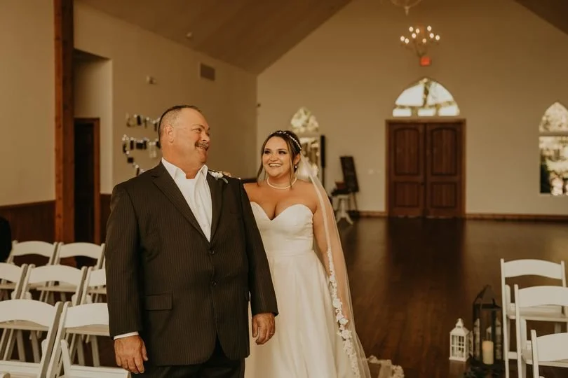 A bride in a white wedding dress and a man in a dark suit stand together inside a decorated wedding venue, smiling at each other.