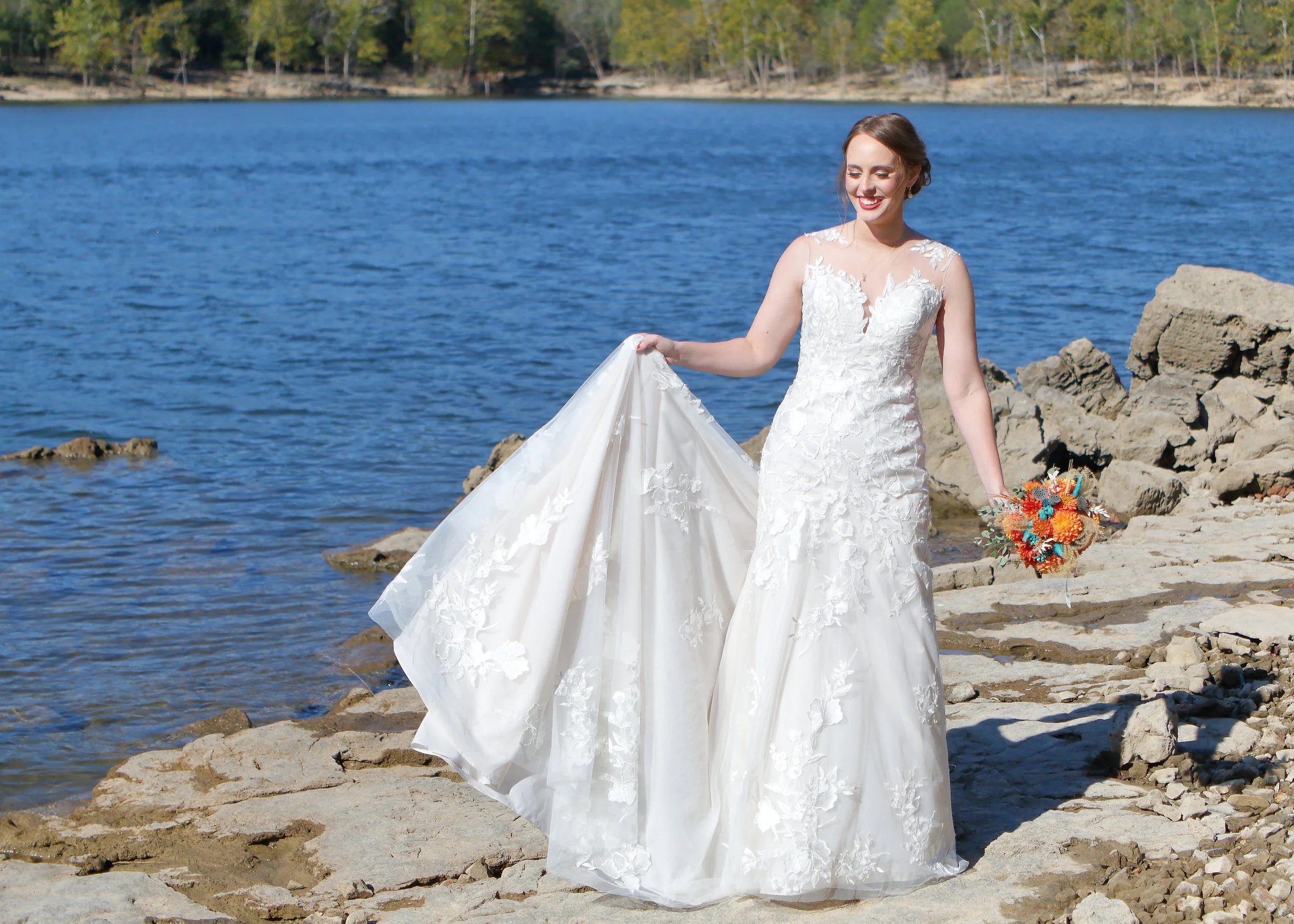 A woman in a white wedding dress standing on rocks by a lake with trees in the background.