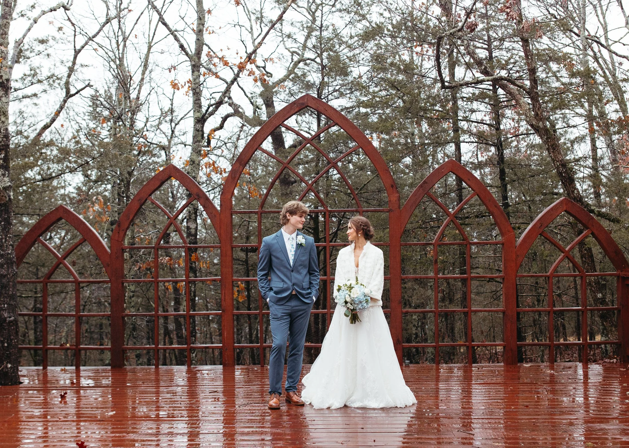 A bride and groom standing together outdoors on a wooden deck during a rainy day, with a rust-colored arched metal structure behind them and trees with leafless branches in the background.