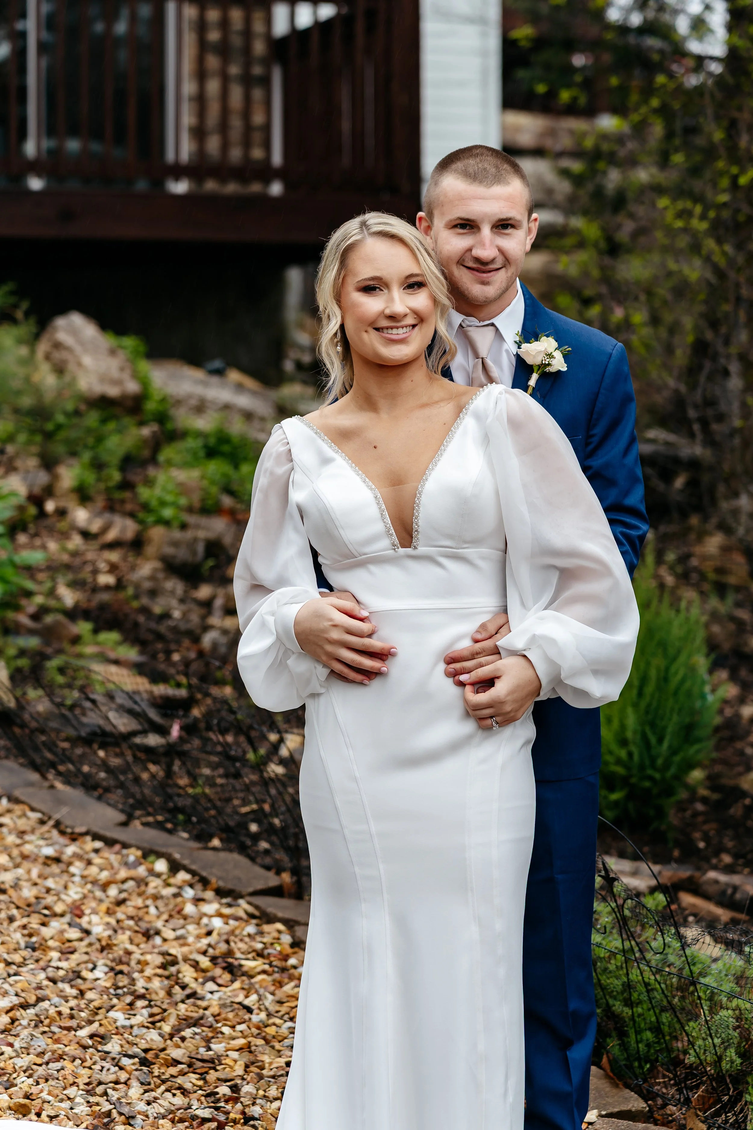 A bride and groom smiling and posing outdoors. The bride wears a white wedding dress with long sleeves and a V-neck, and the groom wears a blue suit with a white shirt and beige tie. The groom has a white boutonniere on his lapel, and they are standi