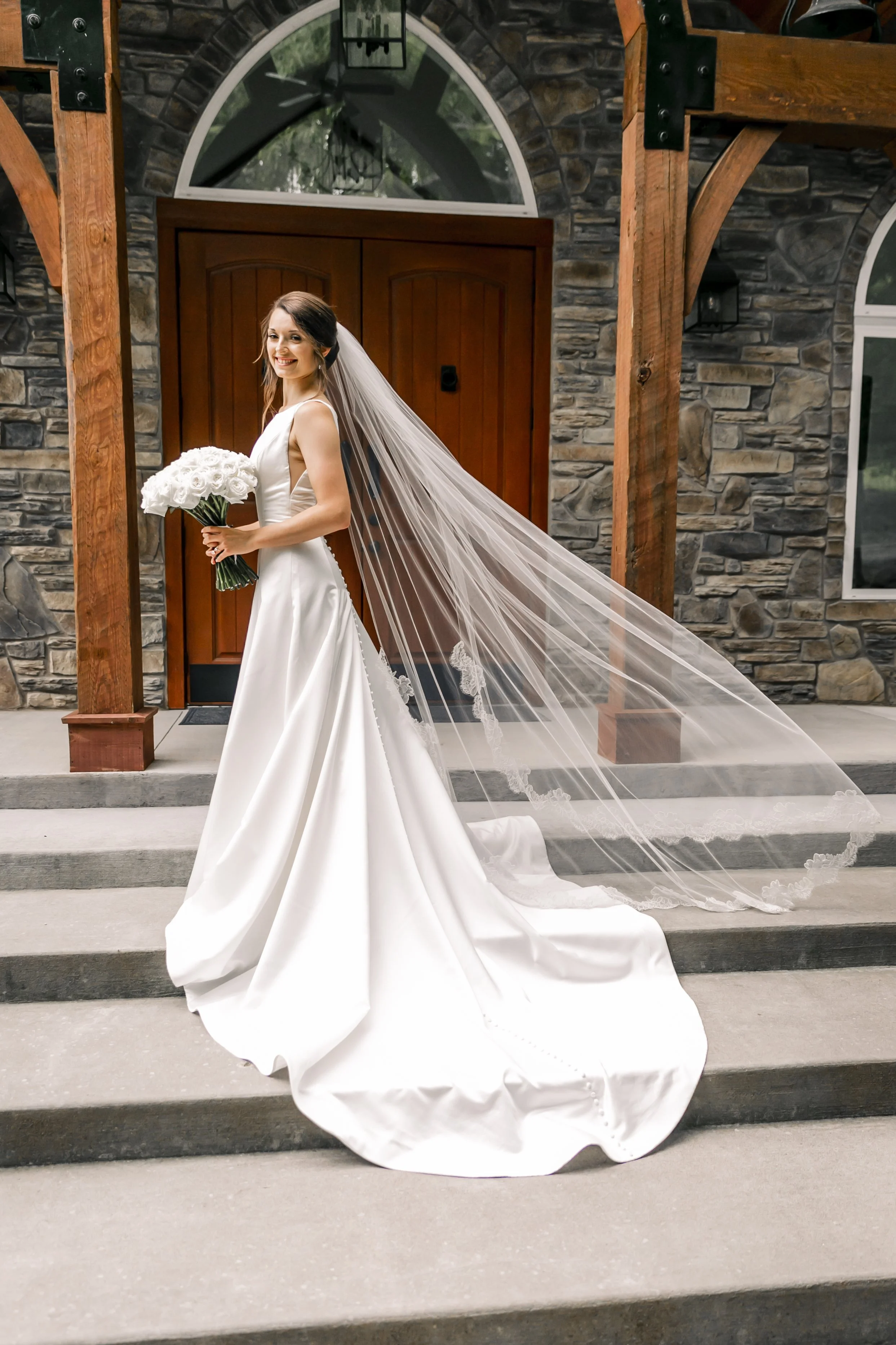 Bride holding a bouquet of white roses, standing on steps outside a stone building with wooden door and arched window above, wearing a white wedding gown and veil.