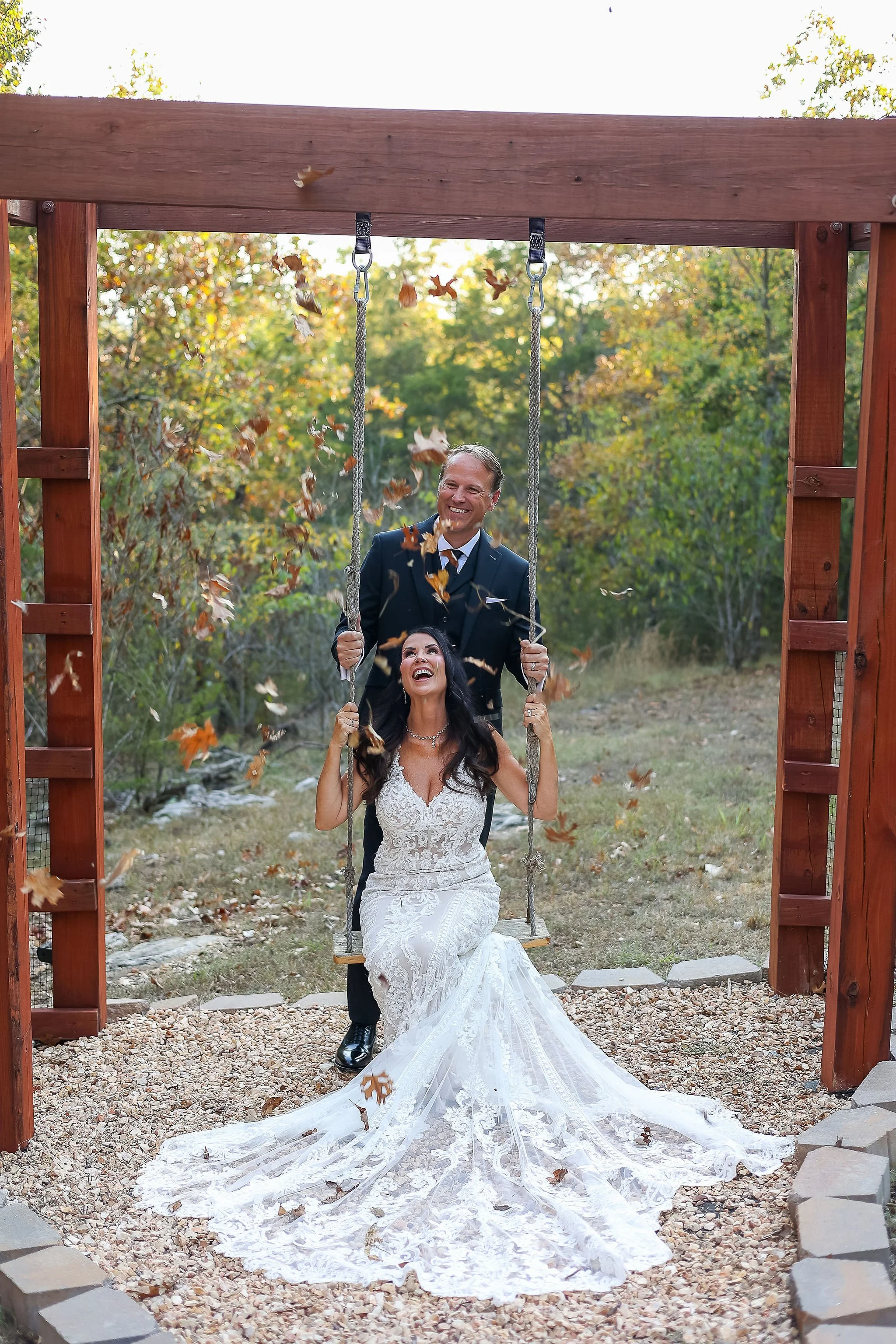 A bride in a white lace wedding gown sitting on a swing, with a groom standing behind her, smiling and holding the swing ropes, outdoors with autumn leaves falling.