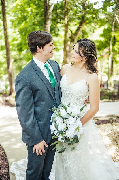 A bride and groom smiling at each other outdoors during their wedding, with trees and sunlight in the background. The bride is holding a white floral bouquet.