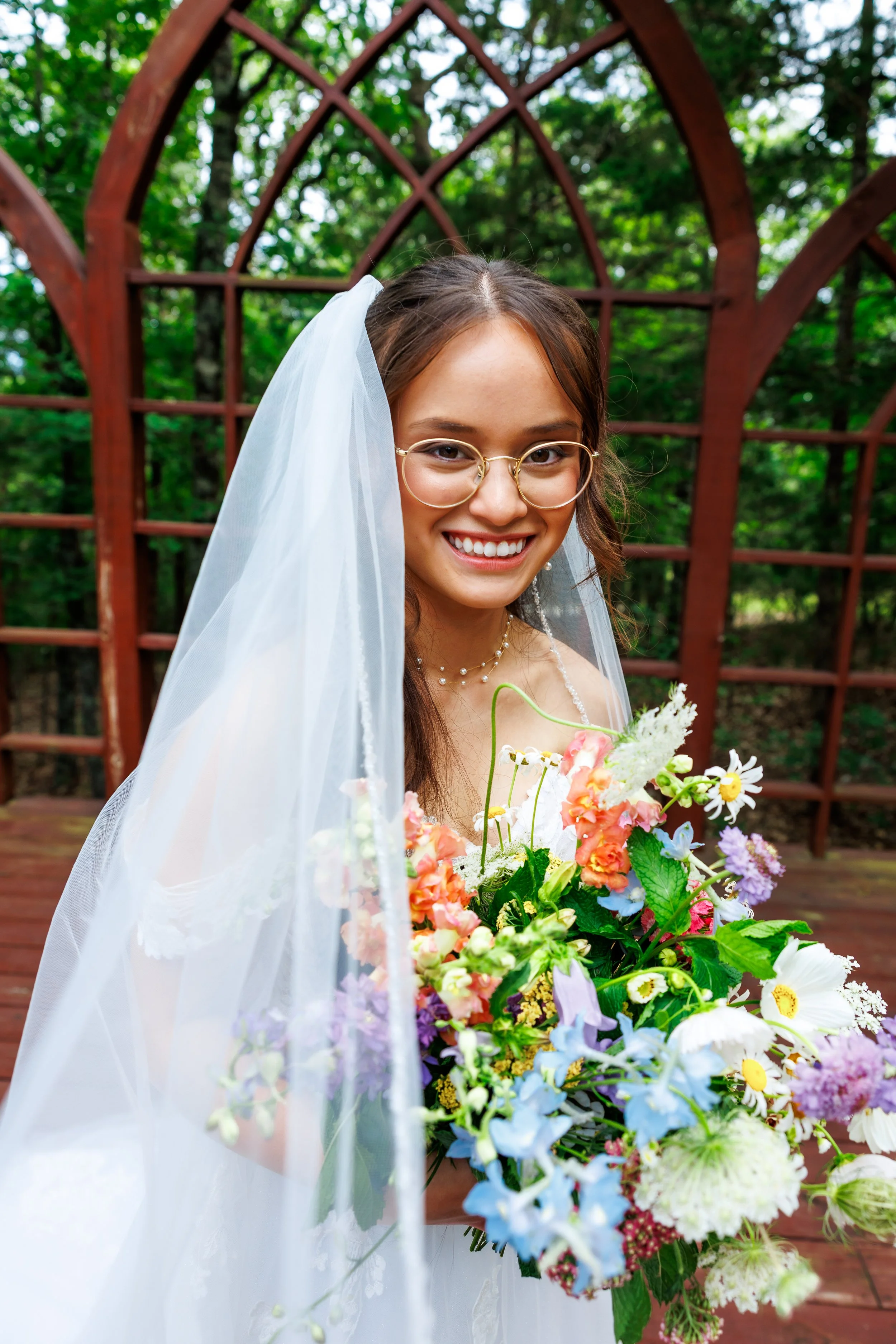 A woman with glasses and a wedding veil holding a large colorful bouquet of flowers outdoors in front of a wooden arch.