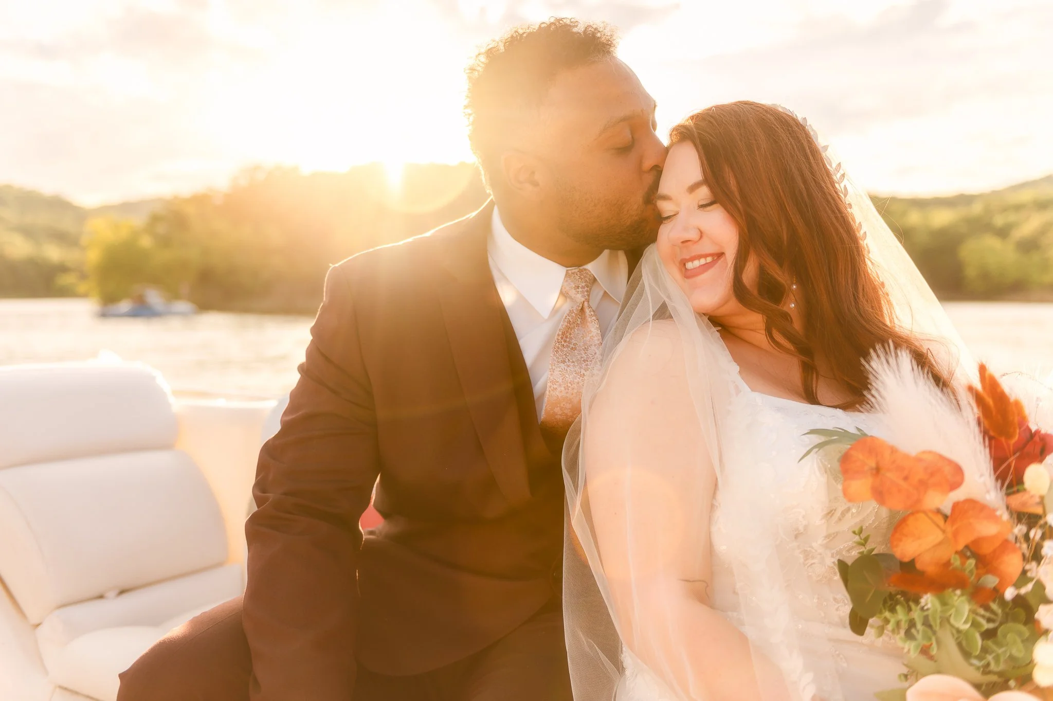 A groom is kissing a bride on the forehead during a wedding ceremony outdoors at sunset, with a blurred background of trees and water.