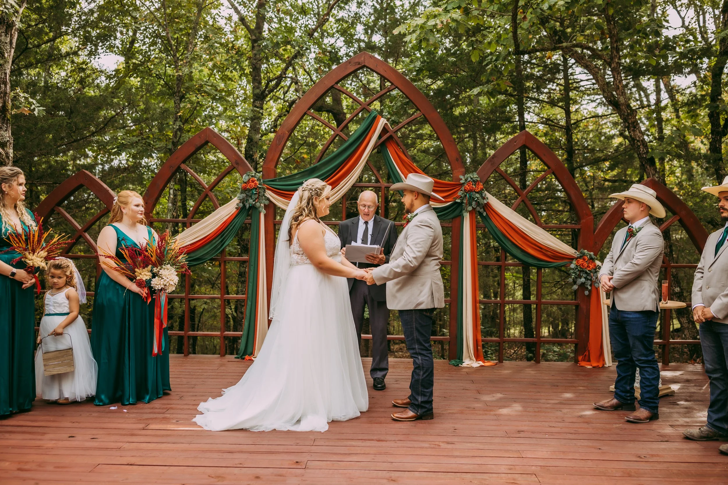 A couple holding hands during their outdoor wedding ceremony in a forest, with an officiant and bridesmaids and groomsmen standing nearby, decorated with floral arrangements and draped fabric on a wooden platform.
