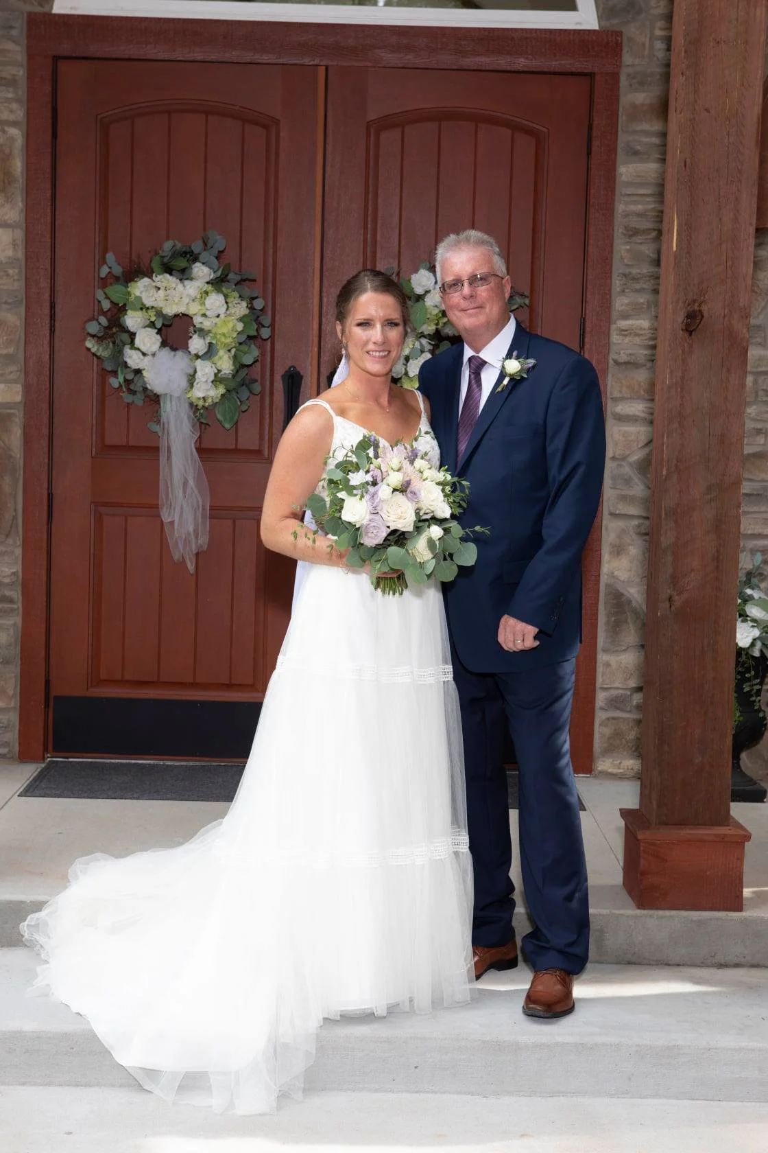 A bride and a man, likely her father, stand in front of a double wooden door decorated with floral arrangements, at a wedding venue. The bride is holding a bouquet, wearing a white wedding dress, and the man is dressed in a navy suit with a boutonnière.