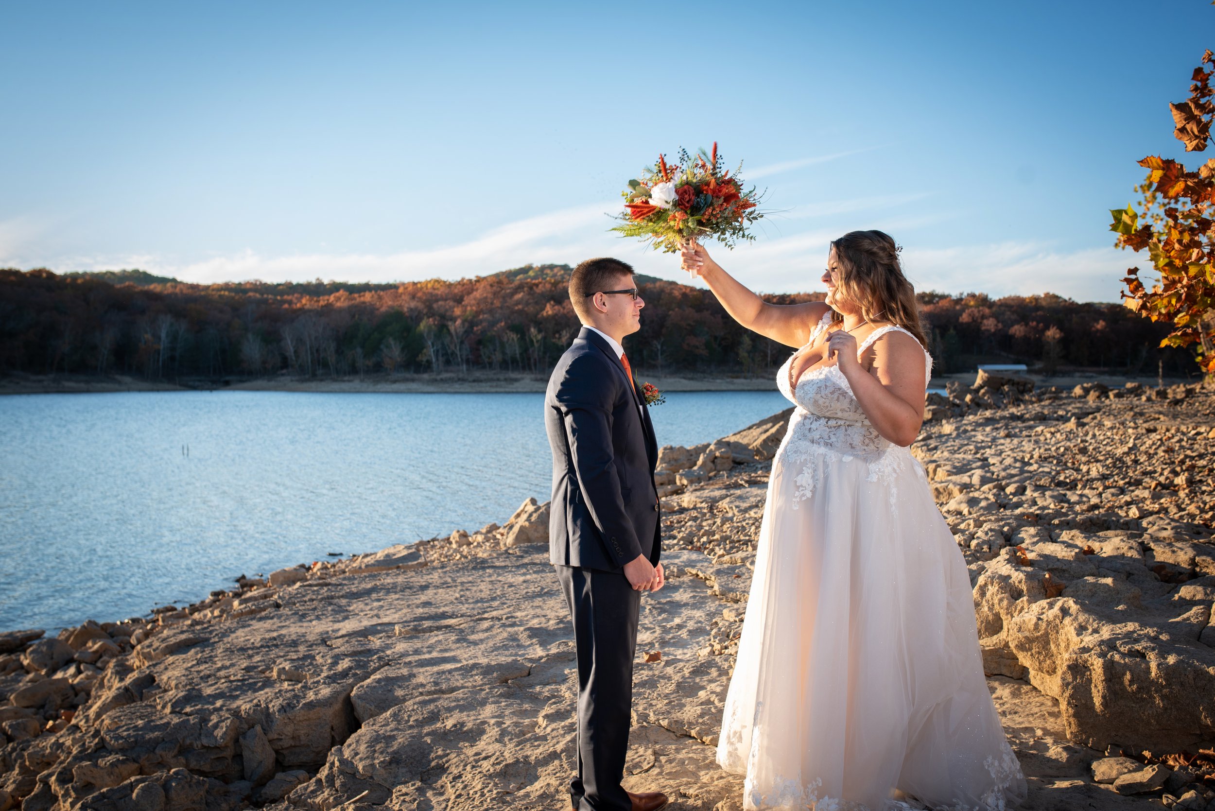 A bride in a white wedding dress and a groom in a suit standing by a lake on a rocky shoreline during sunset, with the bride holding a bouquet of flowers over the groom's head.