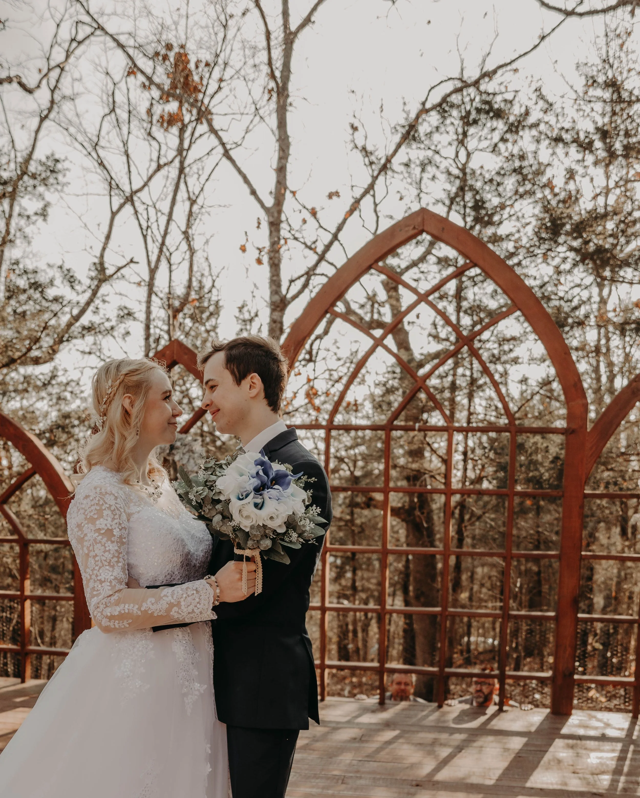A bride and groom stand close together outdoors under an archway, facing each other and smiling. The bride wears a white lace wedding gown and holds a bouquet, while the groom is dressed in a dark suit with a boutonniere. The scene is set in a wooded