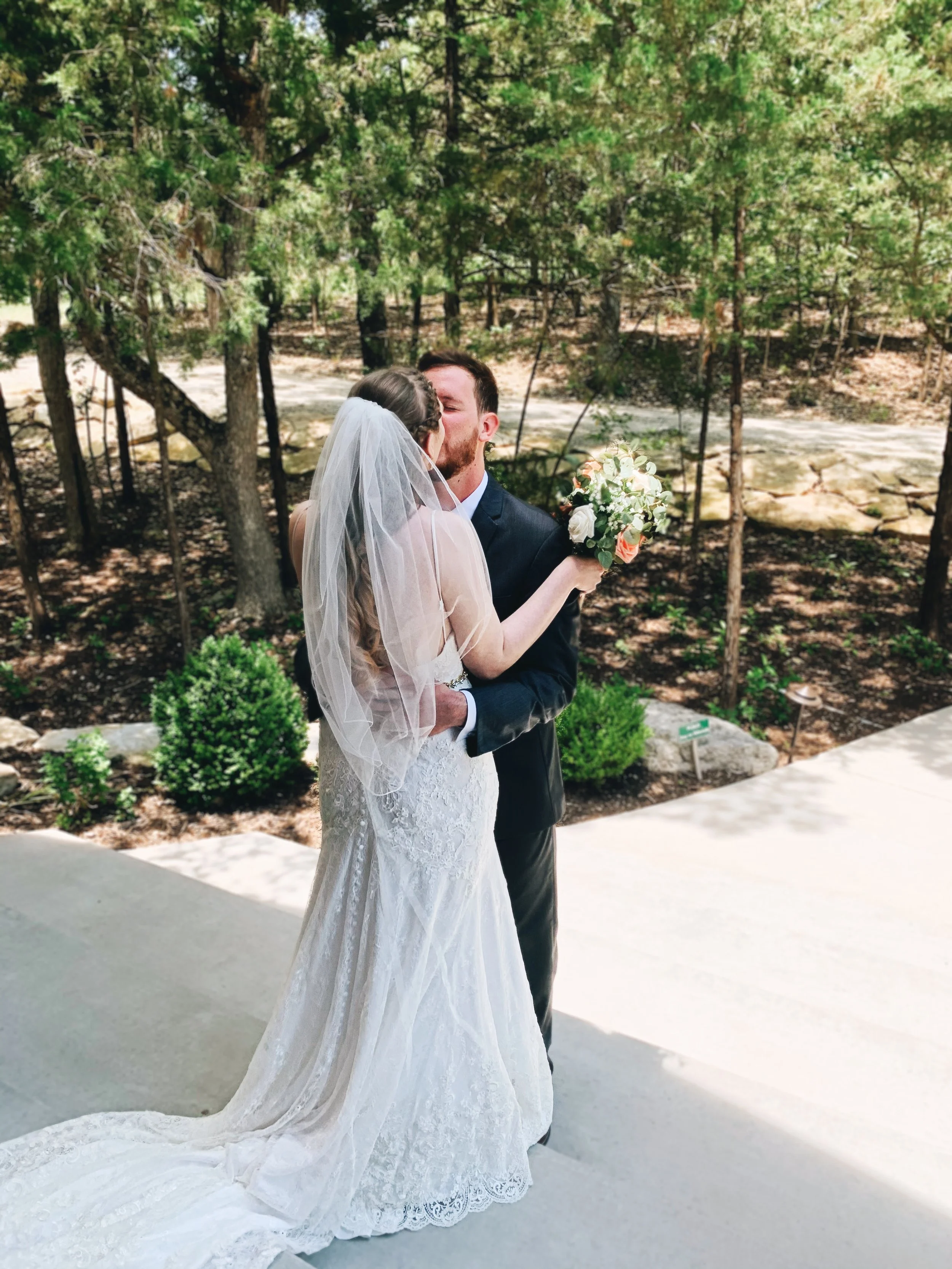 A bride and groom sharing a kiss outdoors during a wedding ceremony or photoshoot, with the bride wearing a lace wedding gown and veil, and the groom in a dark suit, surrounded by trees and greenery.