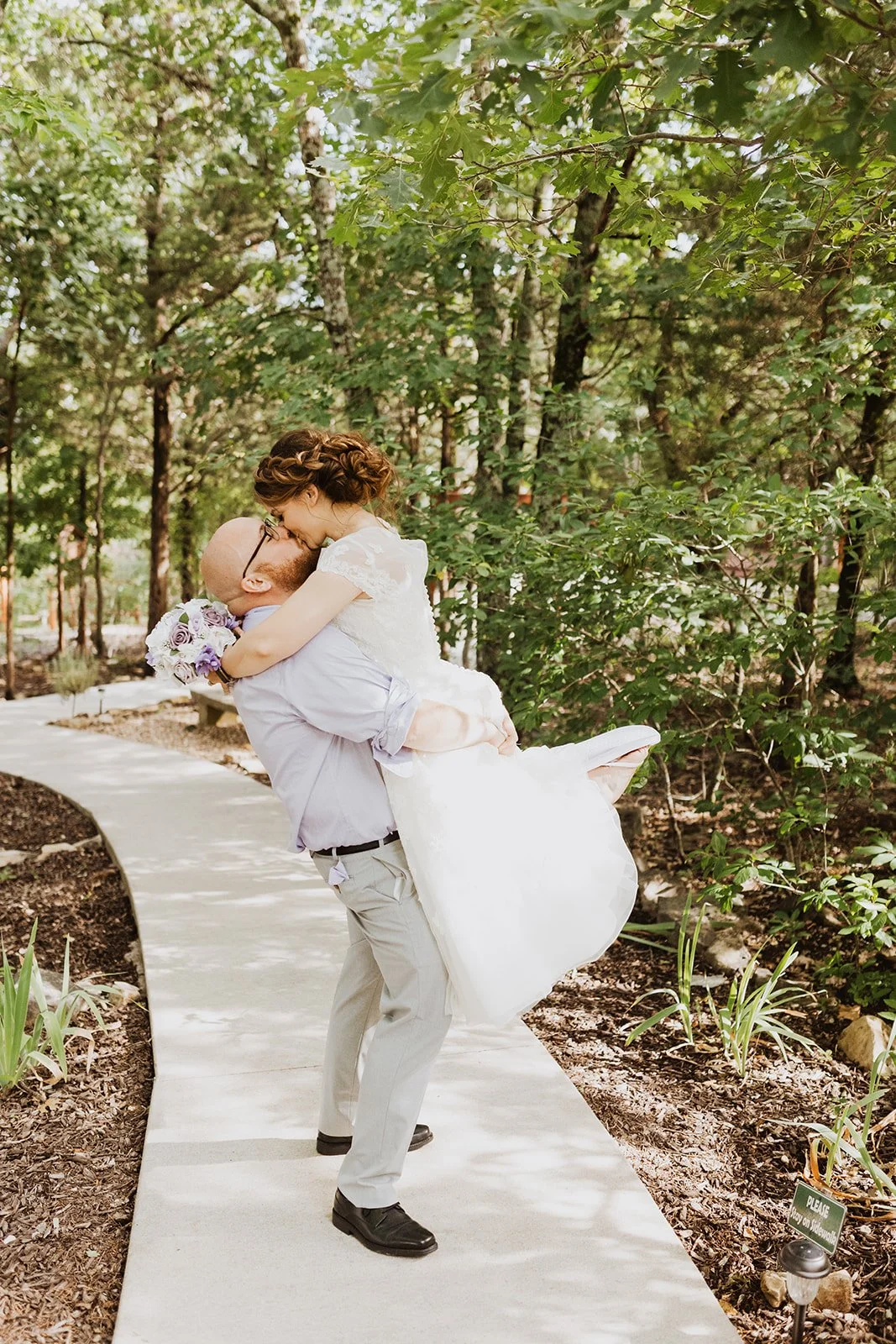 A man in a light dress shirt and trousers is lifting a woman in a white wedding dress and holding a bouquet of purple and white flowers, they are sharing a kiss outdoors on a curved paved walkway surrounded by trees and greenery.