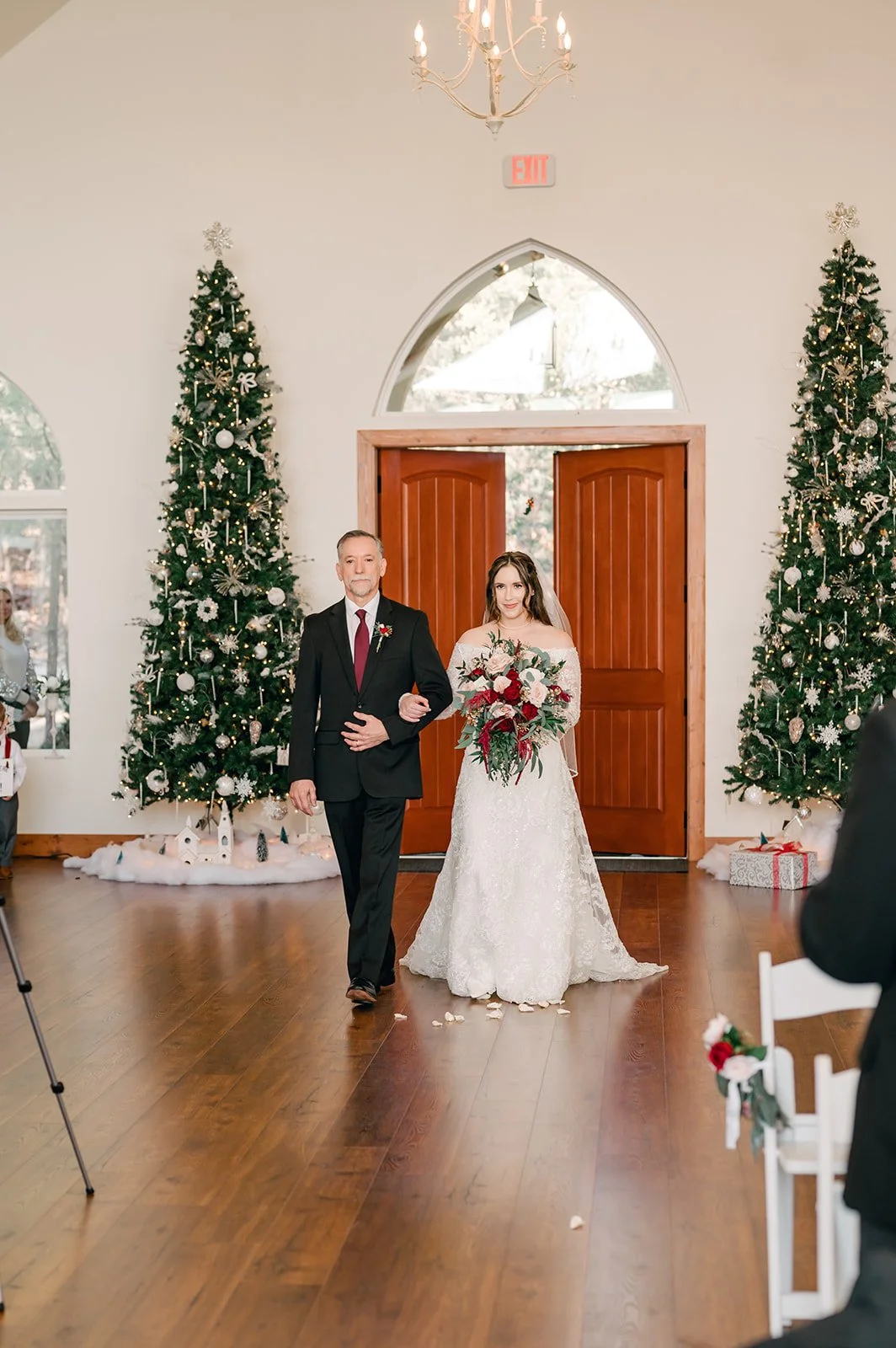 Bride walking down the aisle with her father during a wedding ceremony with Christmas decorations and trees in the background.