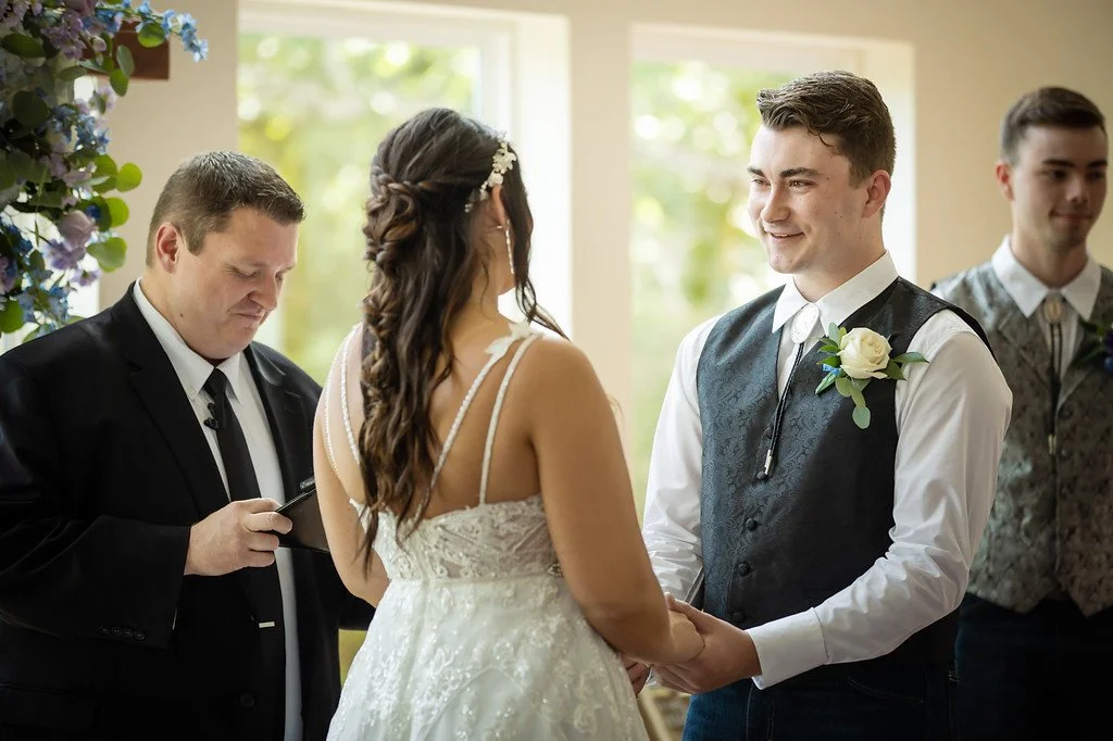 A wedding ceremony with a bride and groom holding hands and smiling at each other, with an officiant reading from a tablet, in a room with large windows and floral decorations.